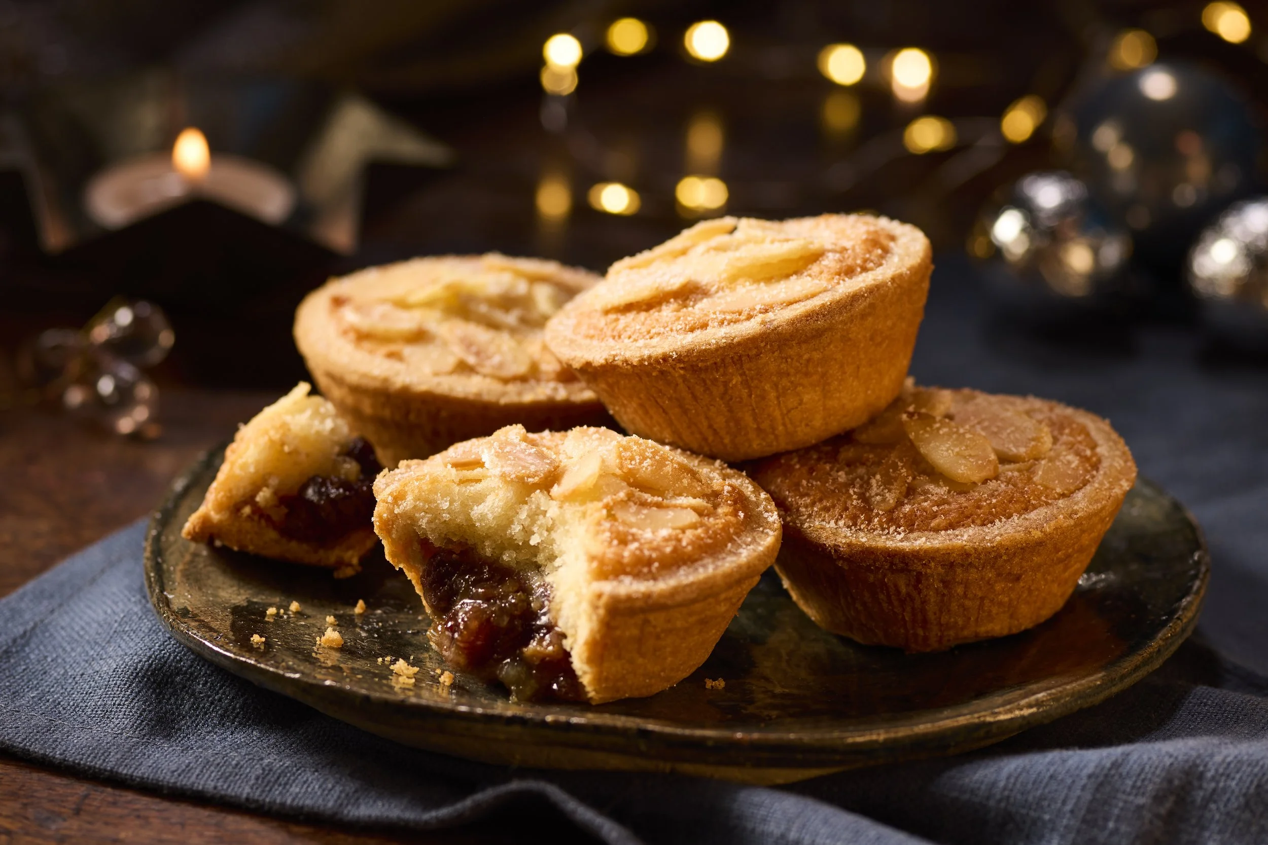 A plate of baked mini apple pies with almond slices on top, some cut open showing apple filling, set on a dark blue cloth with festive decorations in the background.