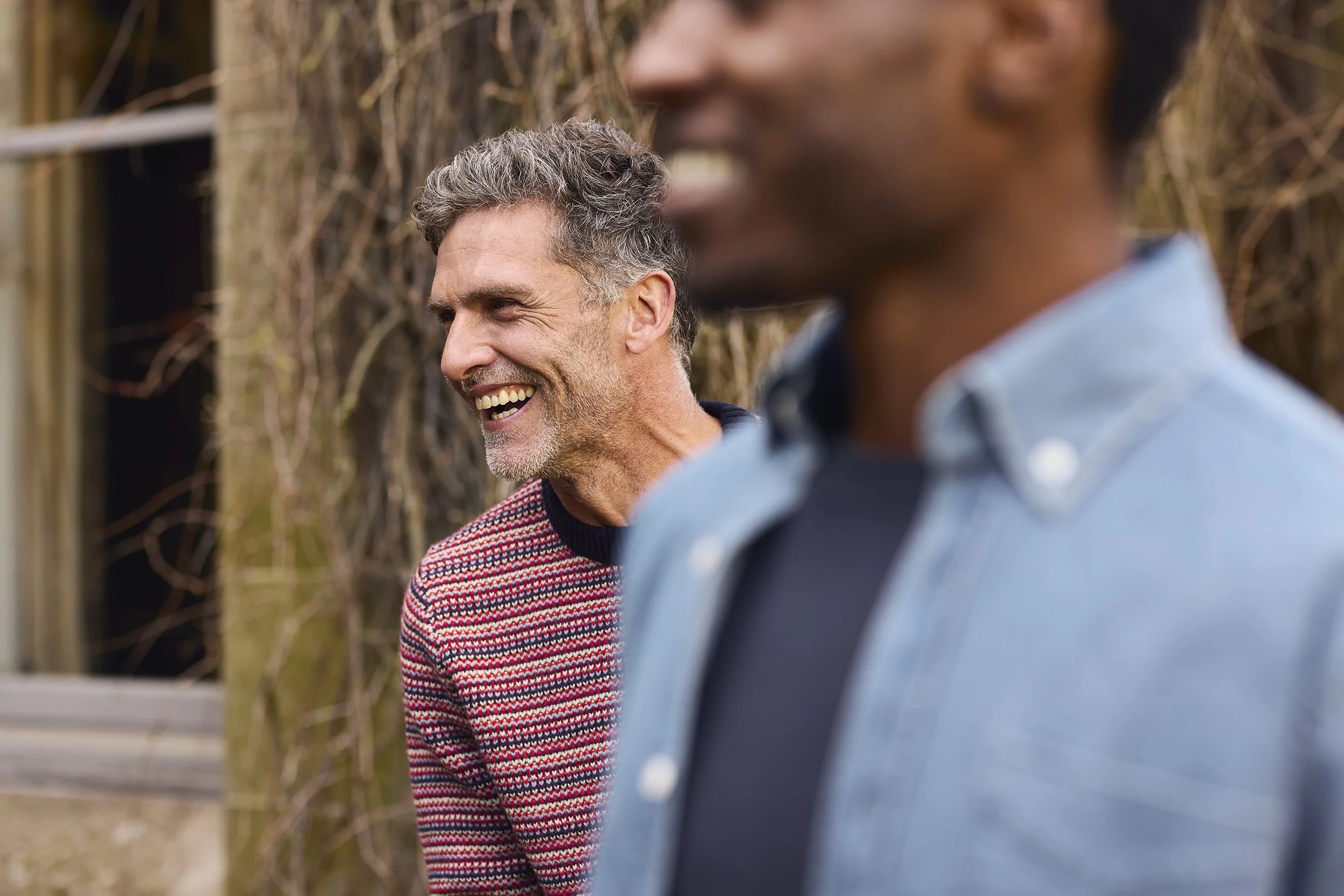 Two men smiling outdoors, one in the background and one in the foreground, with a blurred background of trees and wooden fence.