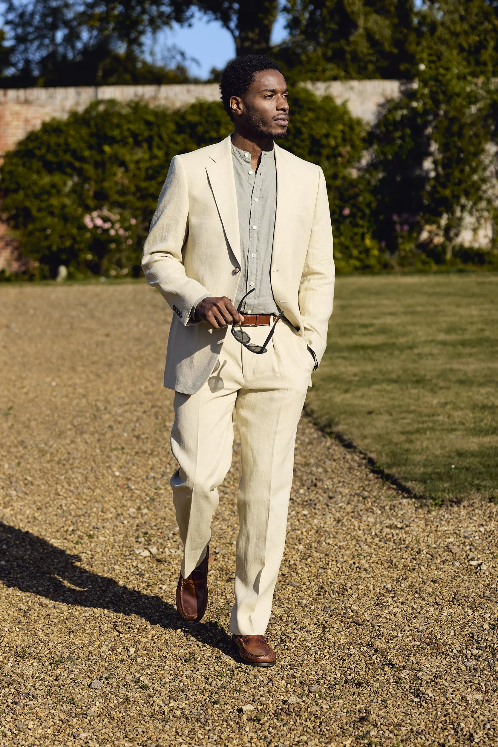 A man walking outdoors on a gravel path, dressed in a light-colored suit, gray shirt, and brown shoes, holding sunglasses in his right hand, with greenery and a brick wall in the background.