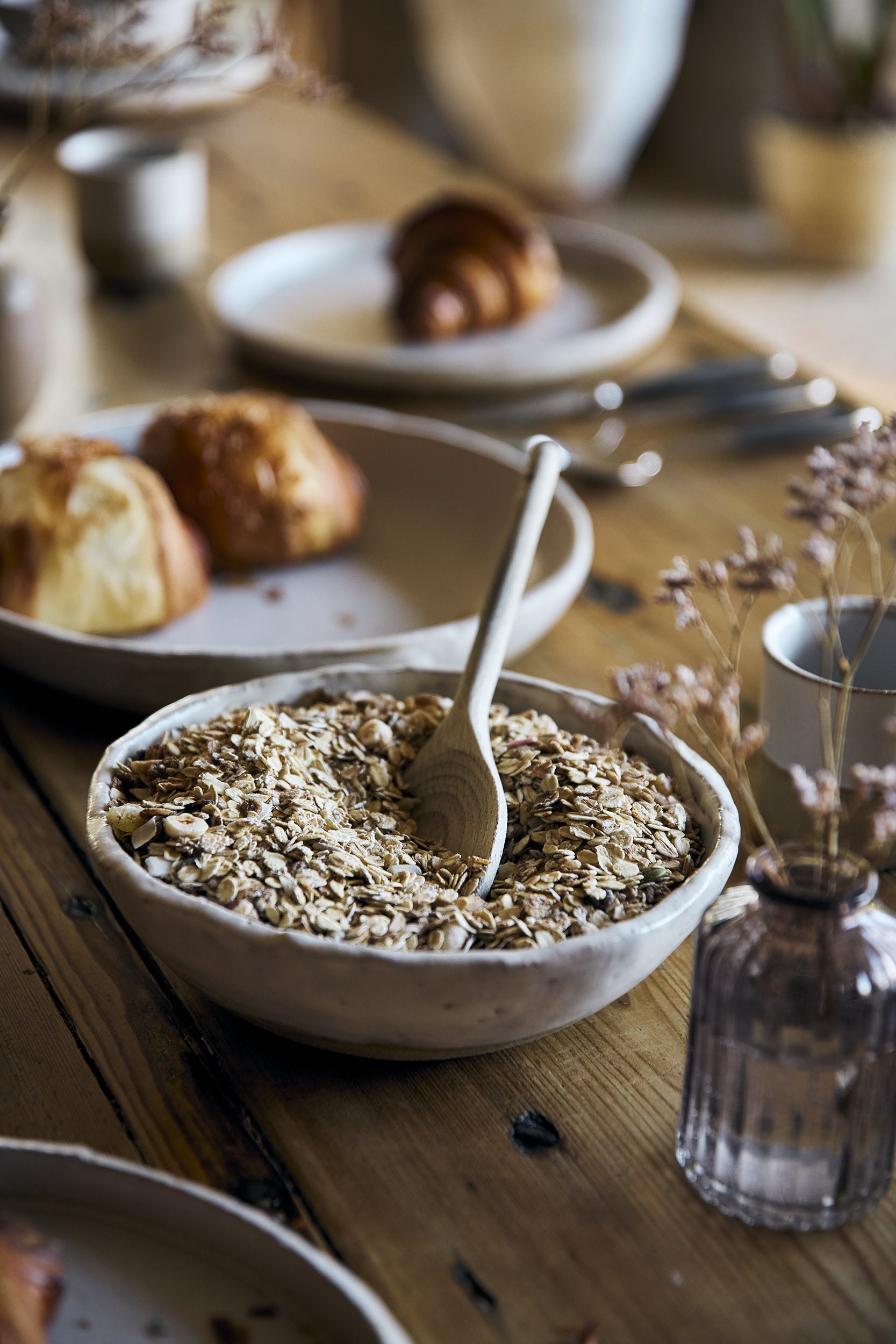 A ceramic bowl filled with oats and a wooden spoon in it on a rustic wooden table, with plates of muffins and a croissant in the background, and some dried flowers and a glass jar nearby.
