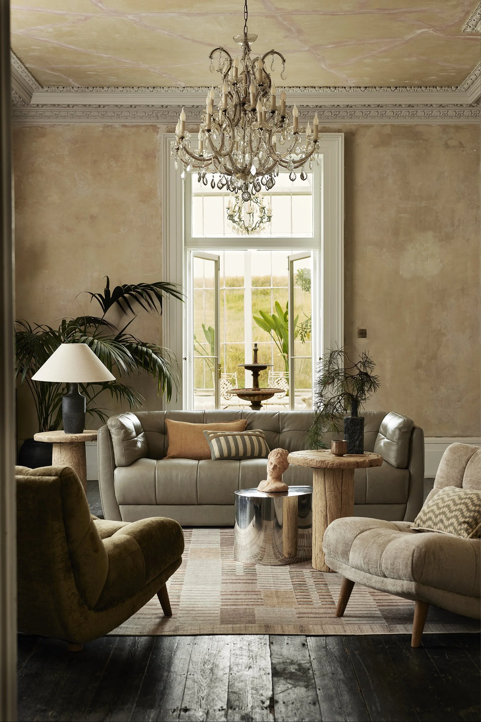 Elegant living room with a chandelier, large window, beige sofa with pillows, armchair, lamps, and decorative plants and sculptures.