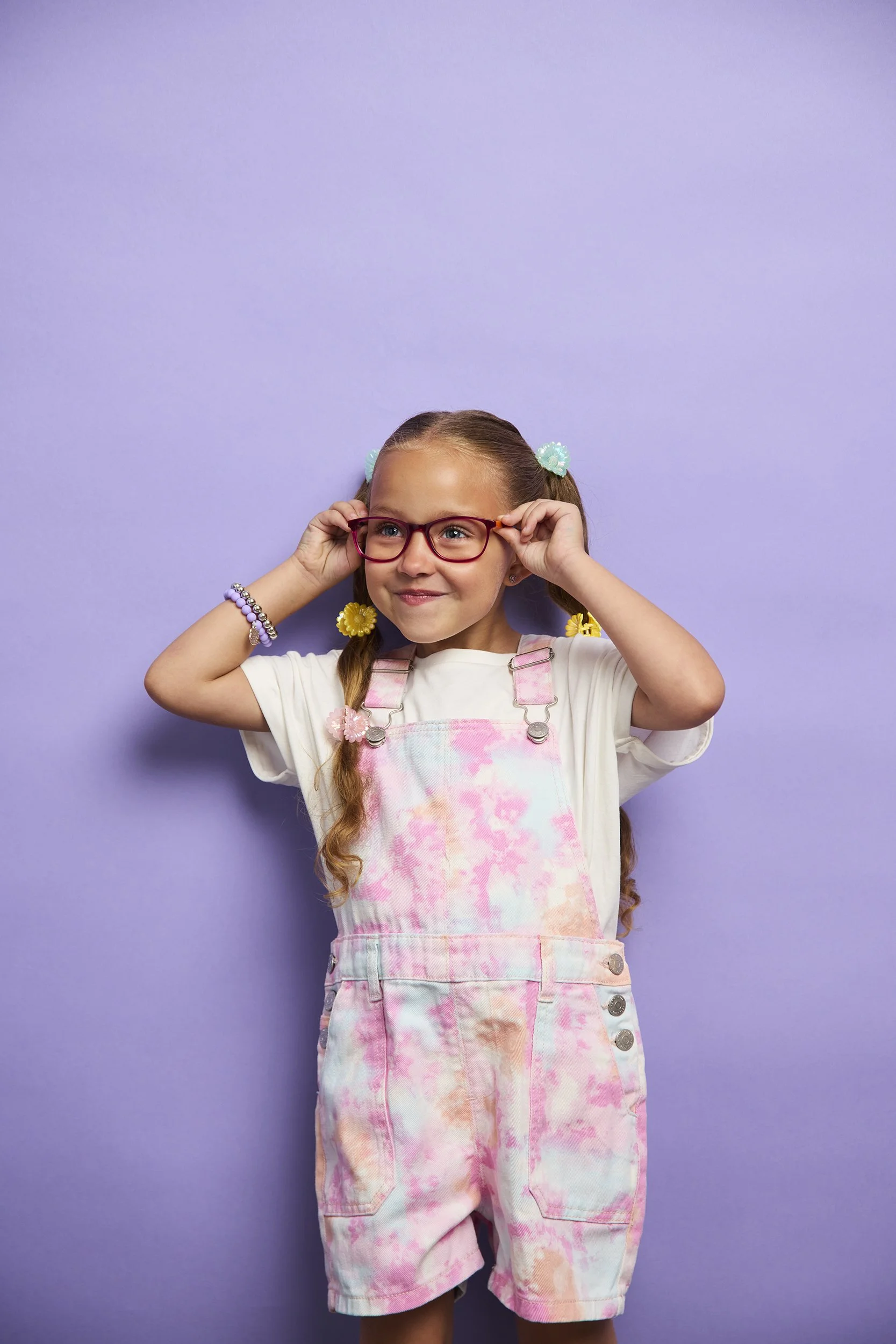 A young girl with glasses, wearing colorful overalls and a white t-shirt, stands in front of a purple background, adjusting her glasses and smiling.