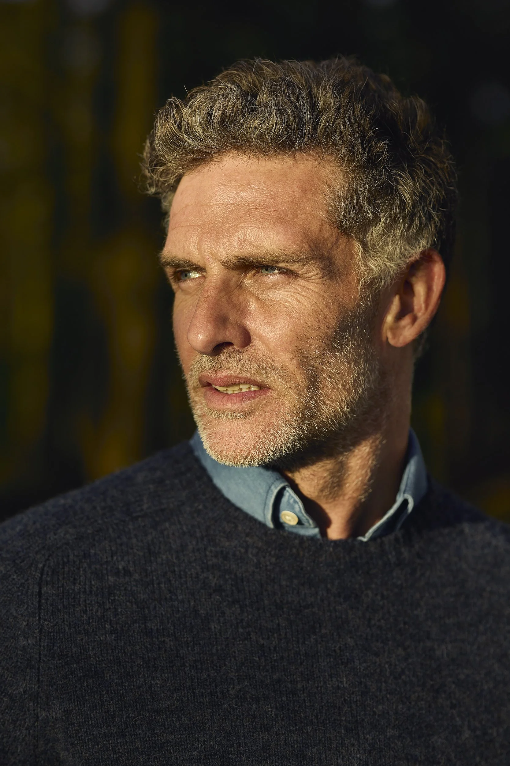 A middle-aged man with gray, wavy hair and a beard, outdoors in a natural setting, looking to the side with sunlight illuminating his face.