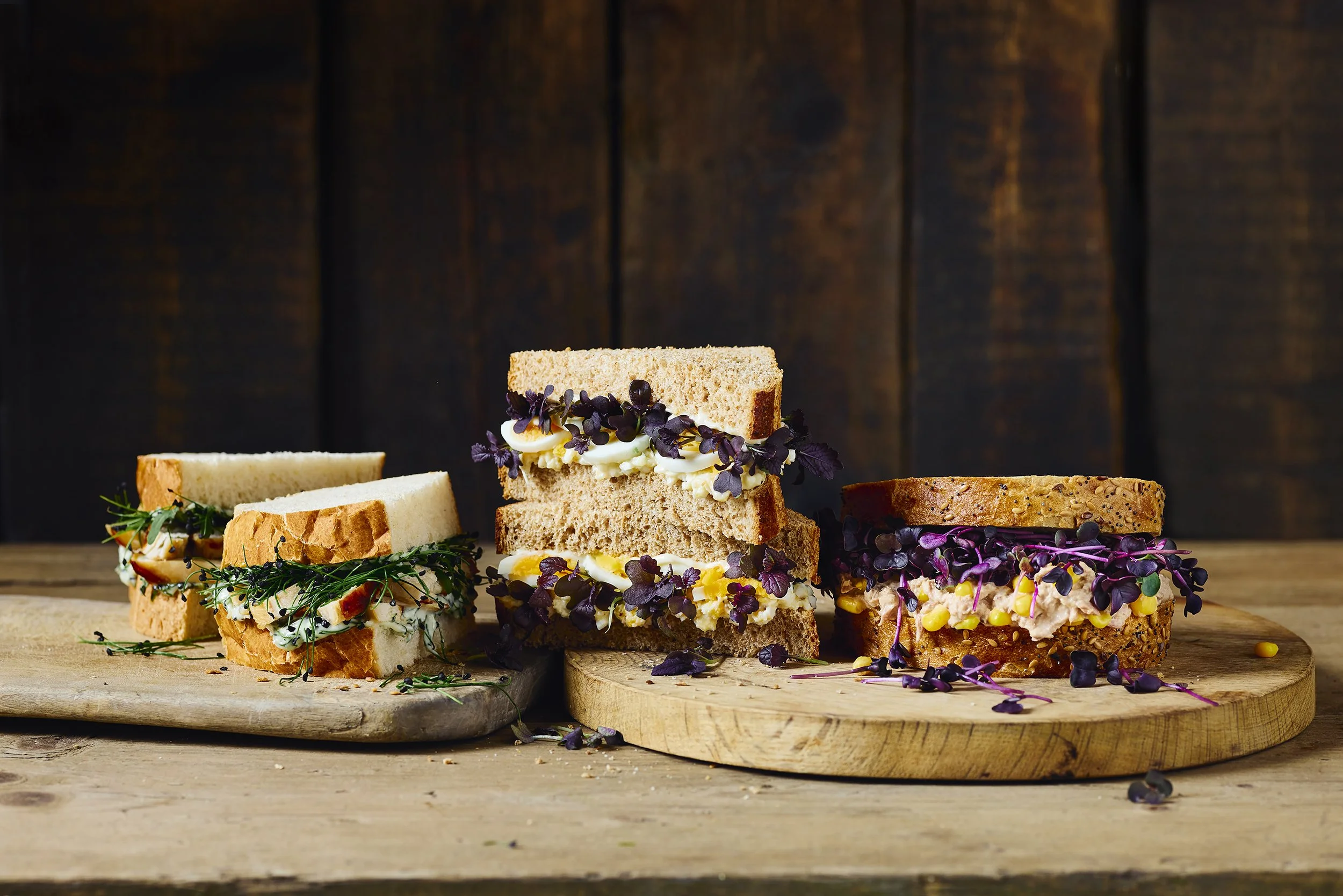 Assorted sandwiches on wooden cutting boards against a dark wood background.