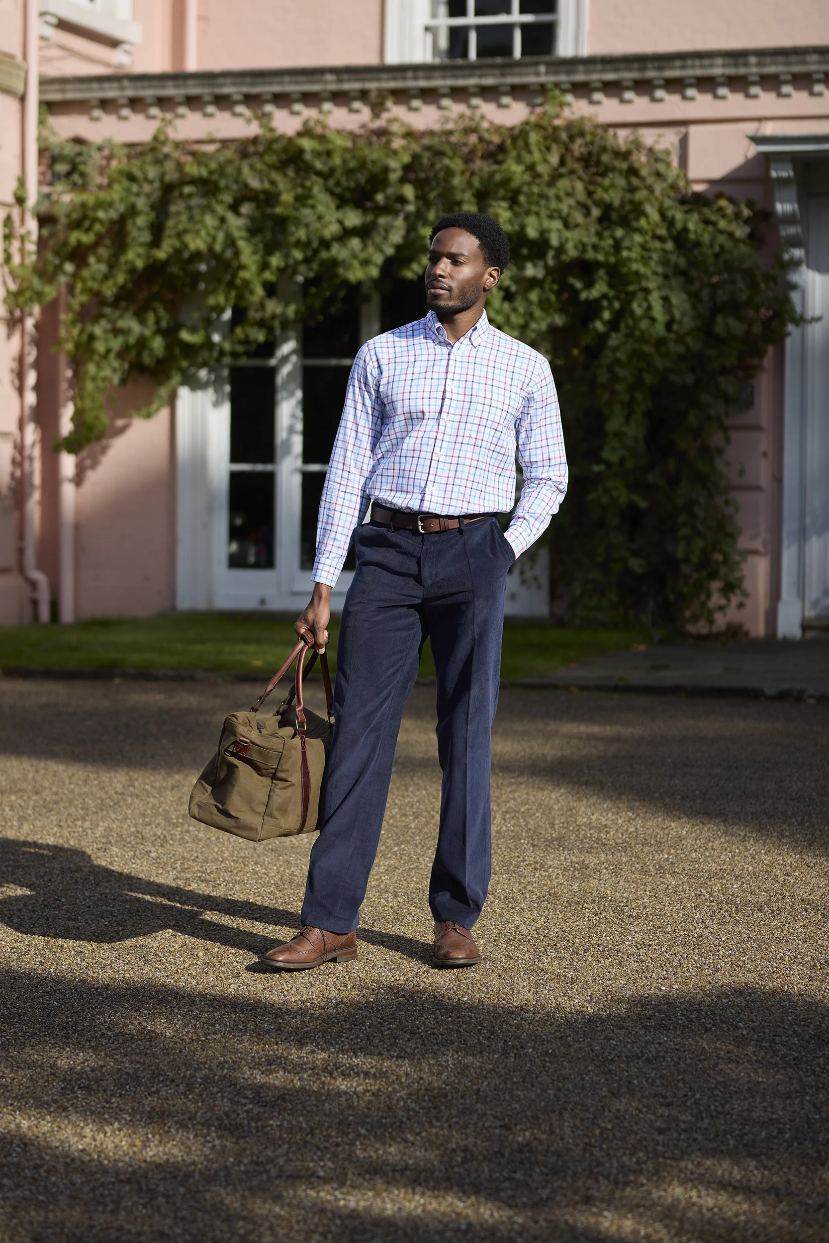 A man in business casual attire holding a briefcase standing outdoors in front of a pink house with greenery.
