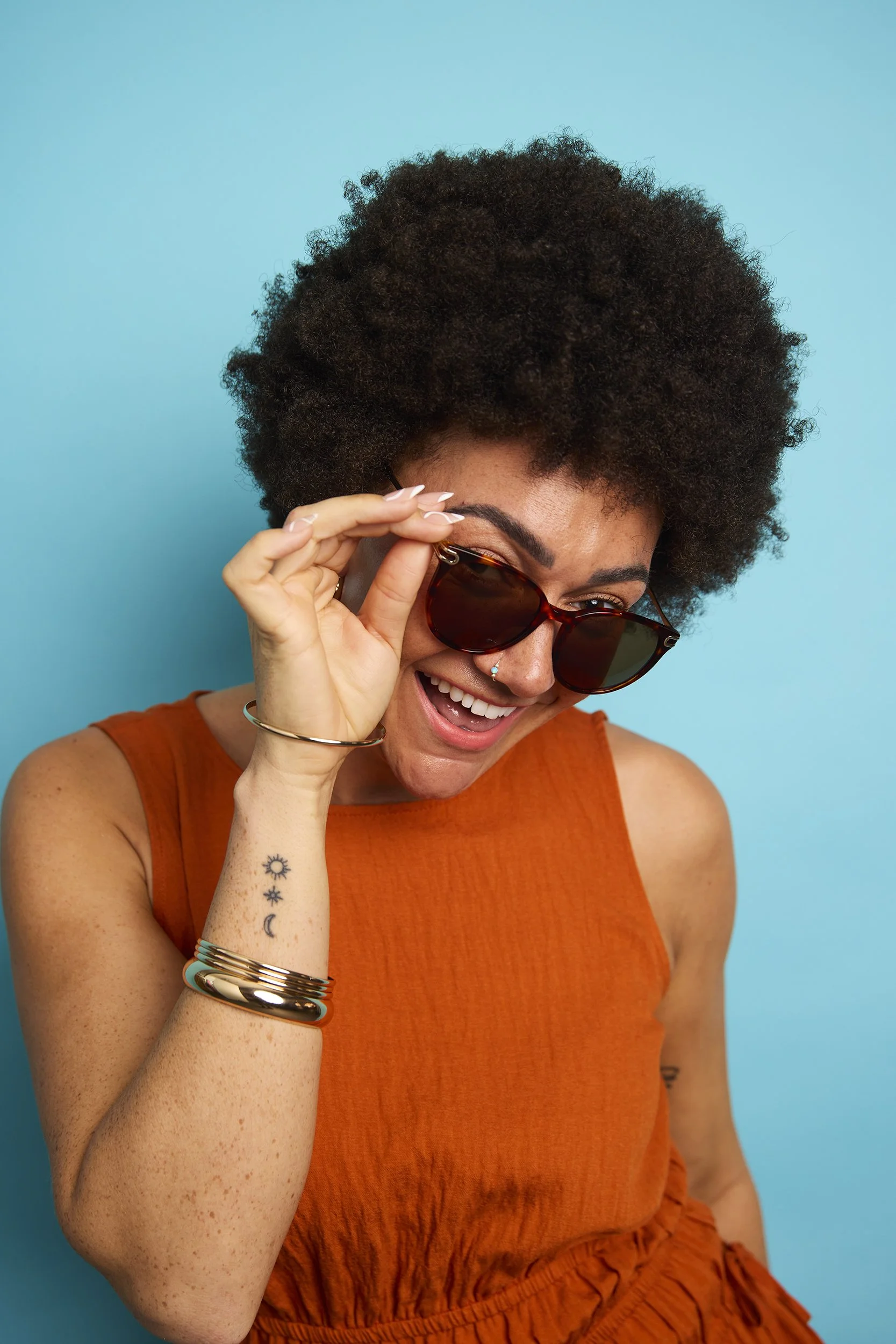 A woman with curly hair wearing sunglasses, orange sleeveless top, and jewelry, smiling against a blue background.
