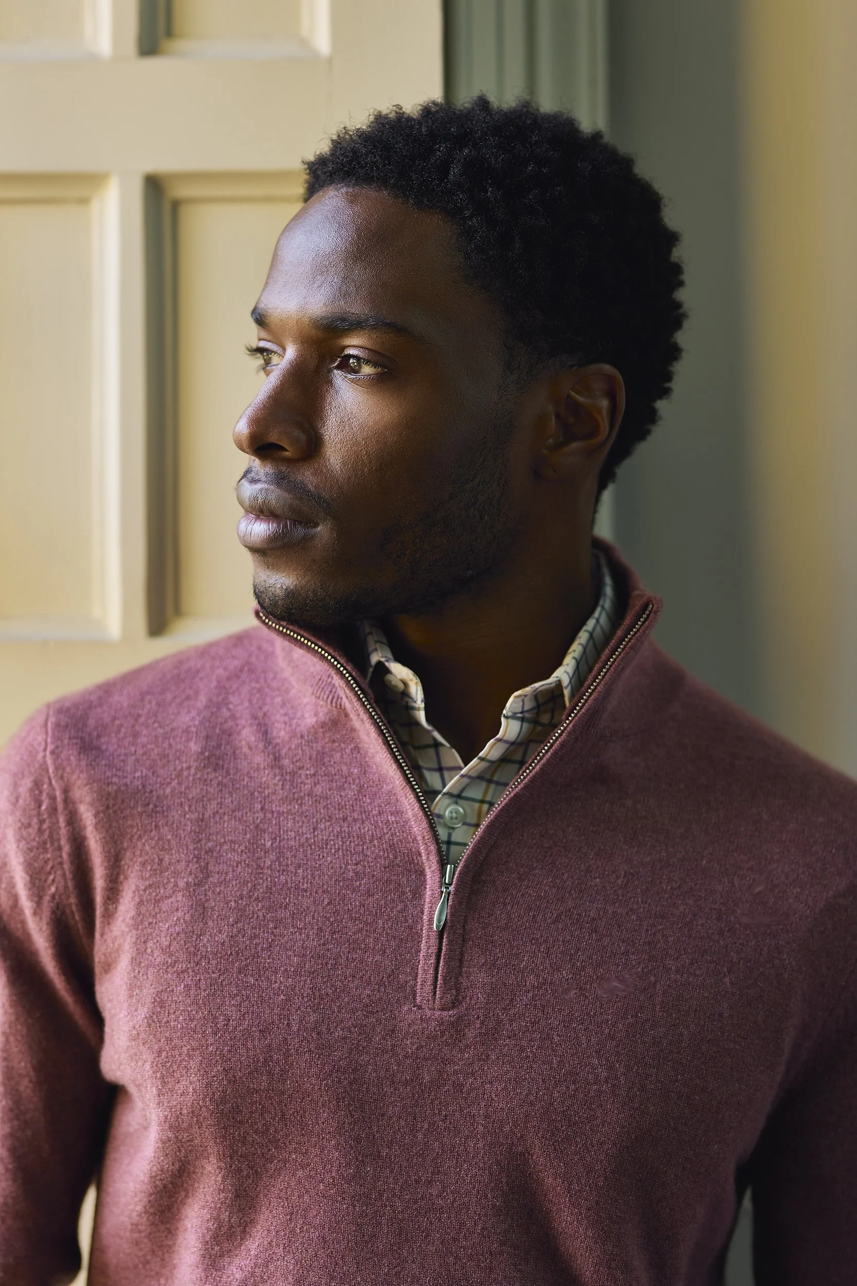 A young African American man looking out a window, wearing a maroon sweater over a collared shirt.