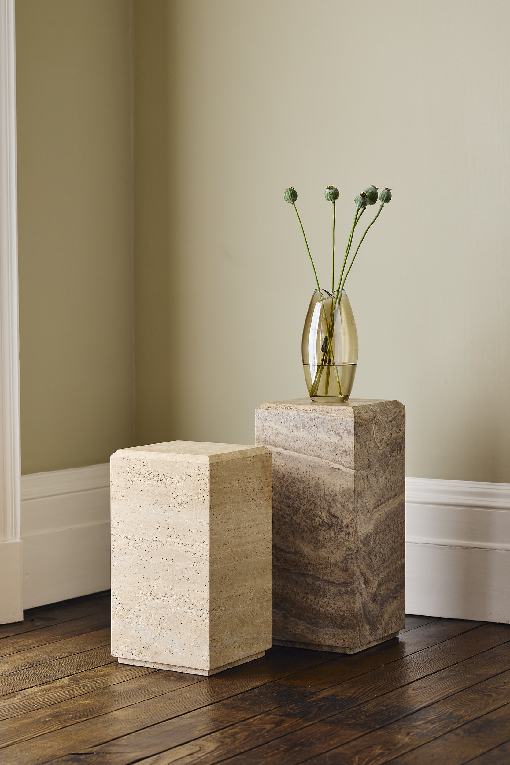 A tall, beige stone pedestal with a smaller, cream-colored stone pedestal beside it, on a dark wooden floor against a light green wall. The taller pedestal has a yellow glass vase with green stems and spherical buds.