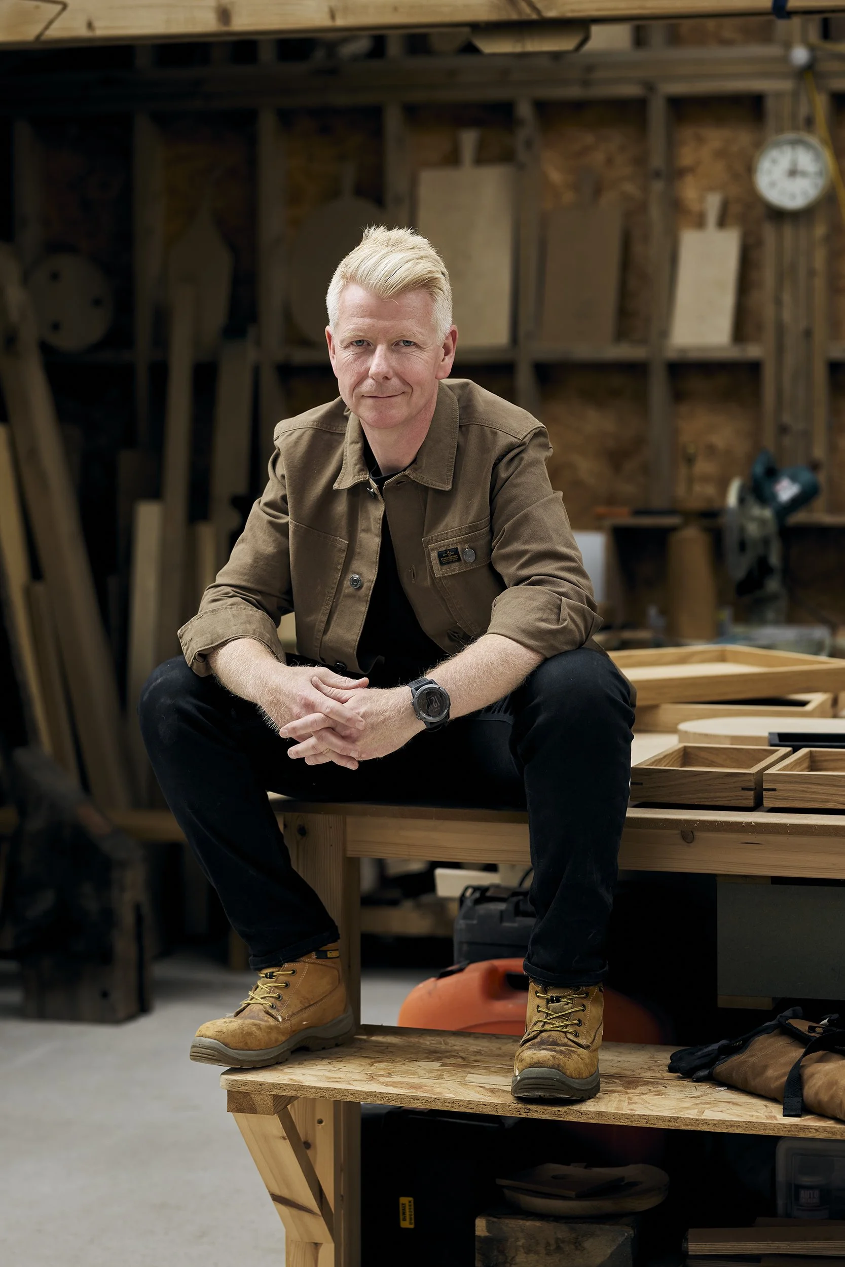 A man in a brown jacket and black pants sitting on a wooden workbench in a woodworking shop, surrounded by woodworking tools and projects.