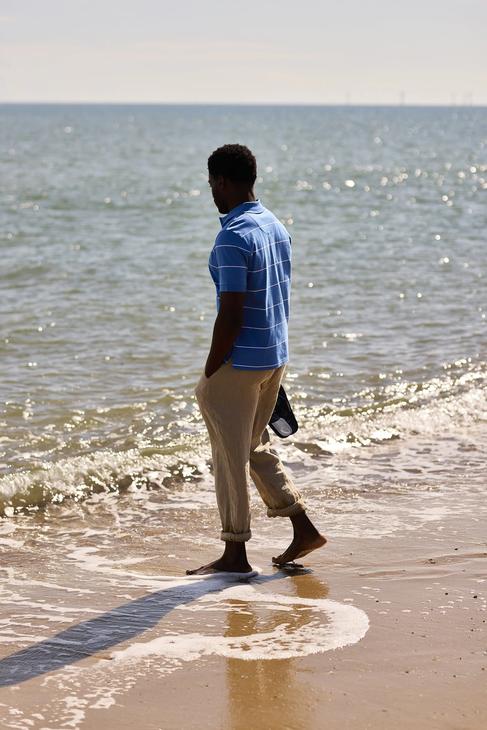 A man in a blue striped shirt and beige pants walking barefoot along the shore of the ocean with his hands in his pockets, looking down at the water, during the daytime.