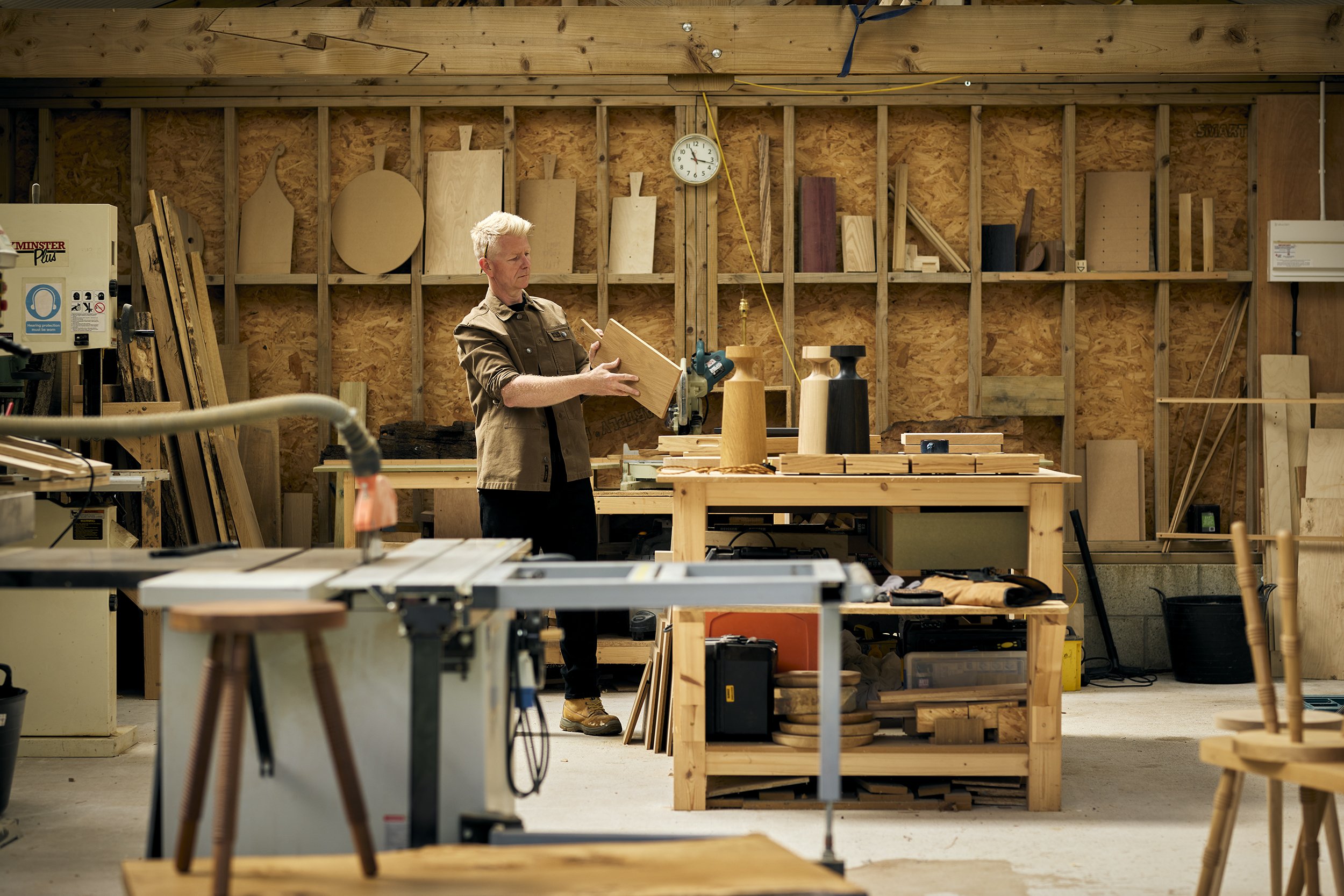 A person in a woodworking shop holding a piece of wood and examining it, surrounded by various woodworking tools and wooden projects.