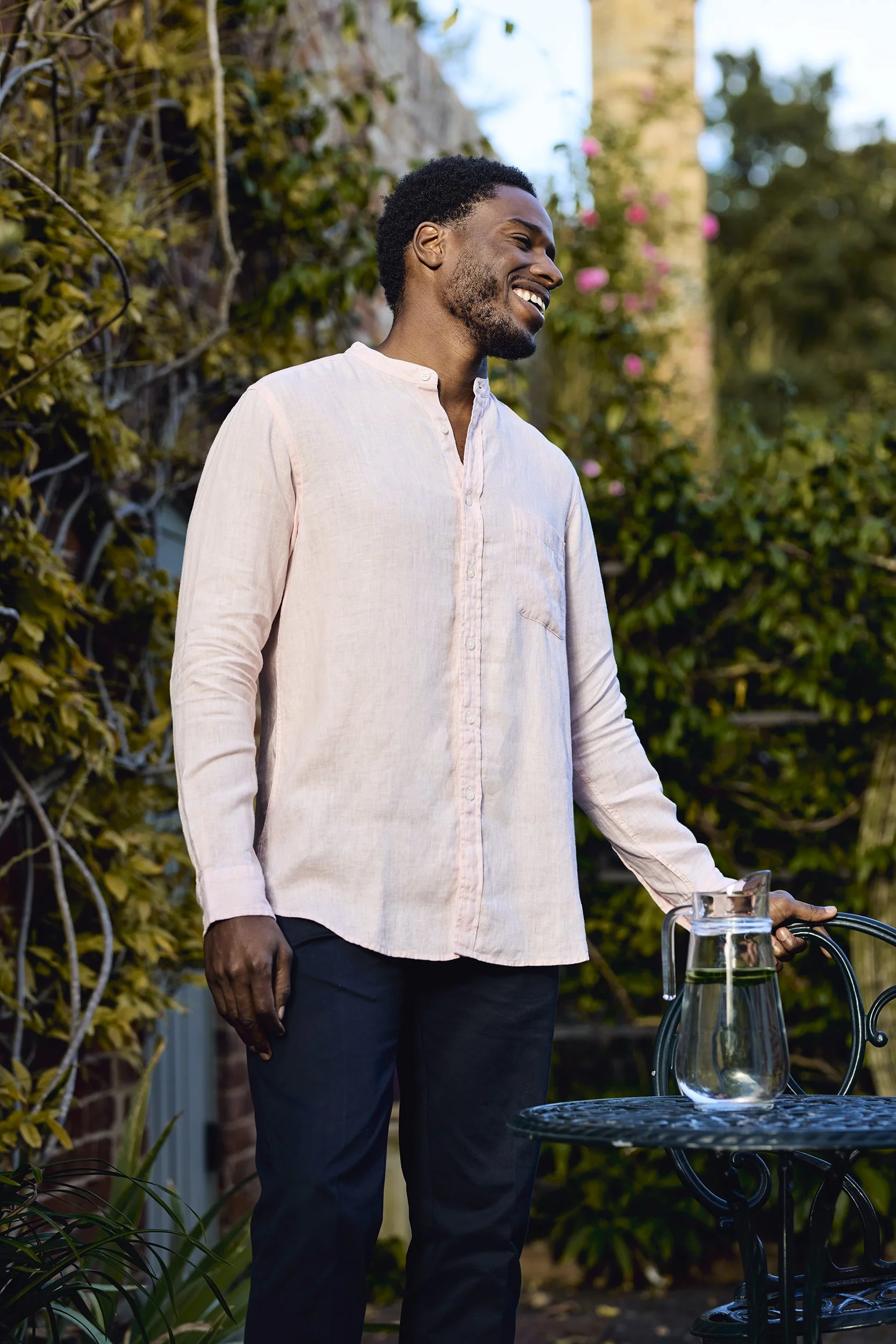A smiling man in a light pink shirt standing outdoors next to a table with a glass pitcher of water, with greenery and flowers in the background.