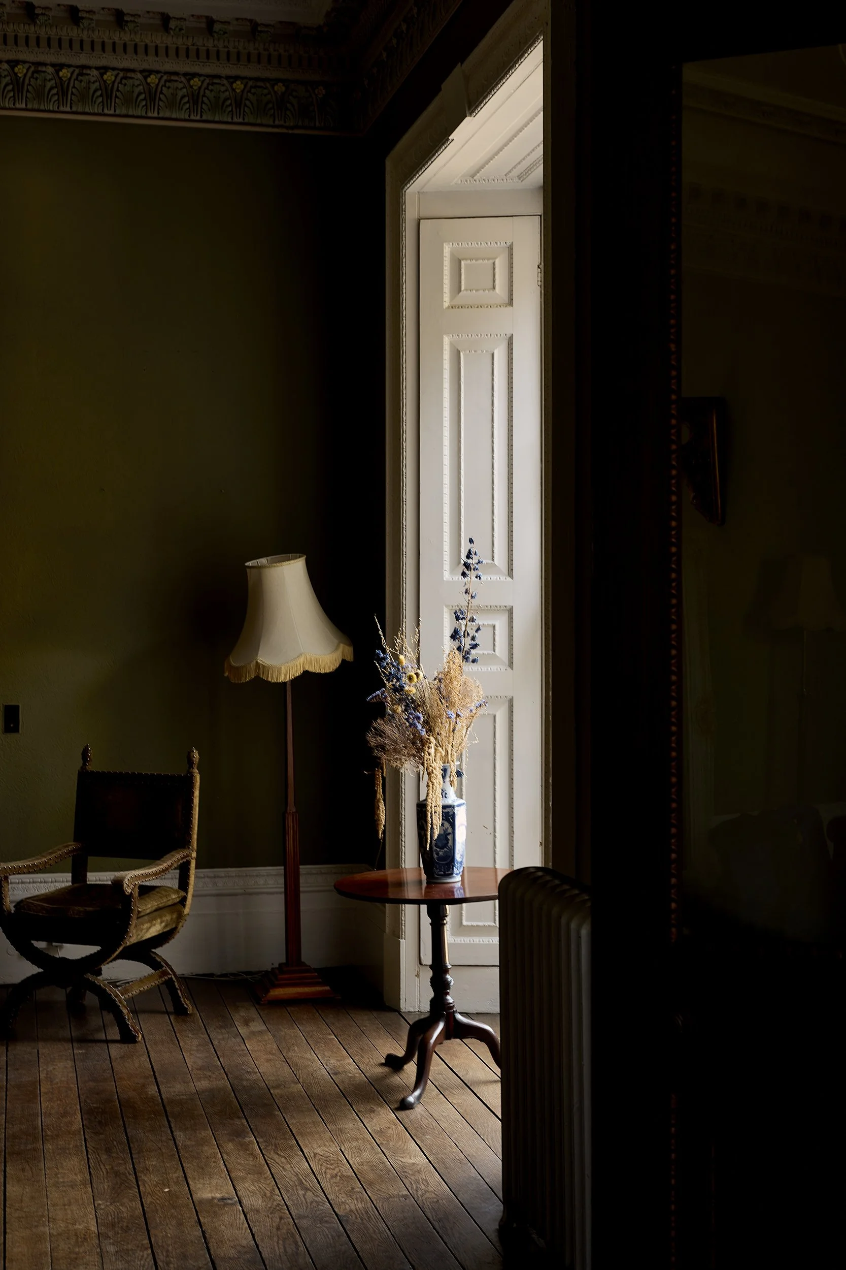 A dimly lit room with wooden floors, an armchair, a standing lamp with a fabric shade, a small round table with a blue and white vase holding dried flowers, near a tall white door with decorative framing, and a dark partial wall or mirror to the righ