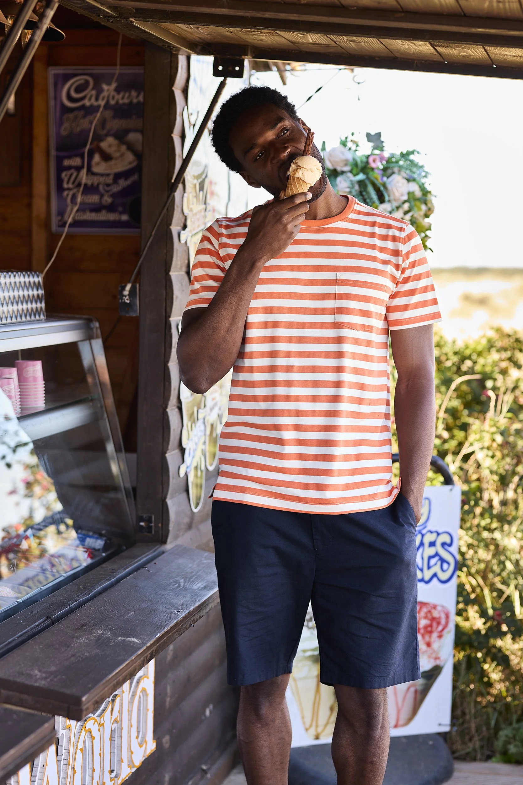 A young man in a red and white striped t-shirt and navy shorts is eating ice cream at an outdoor ice cream stand.