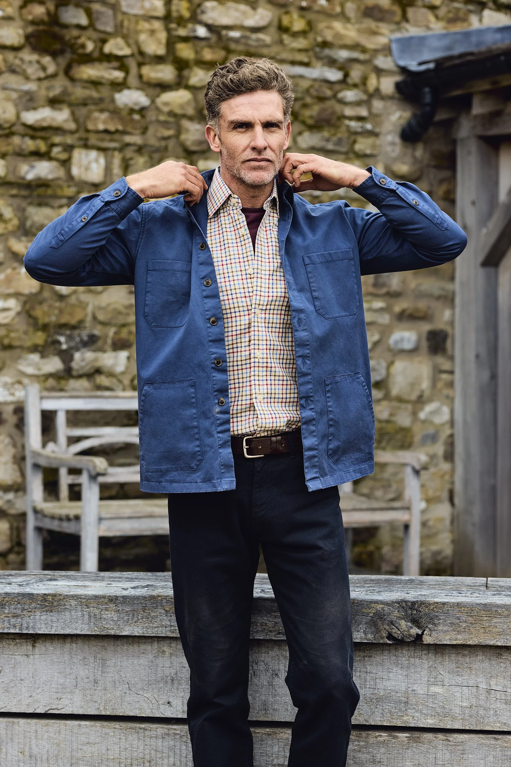 A man in a blue jacket and checkered shirt standing outdoors with a stone wall in the background, adjusting his collar.