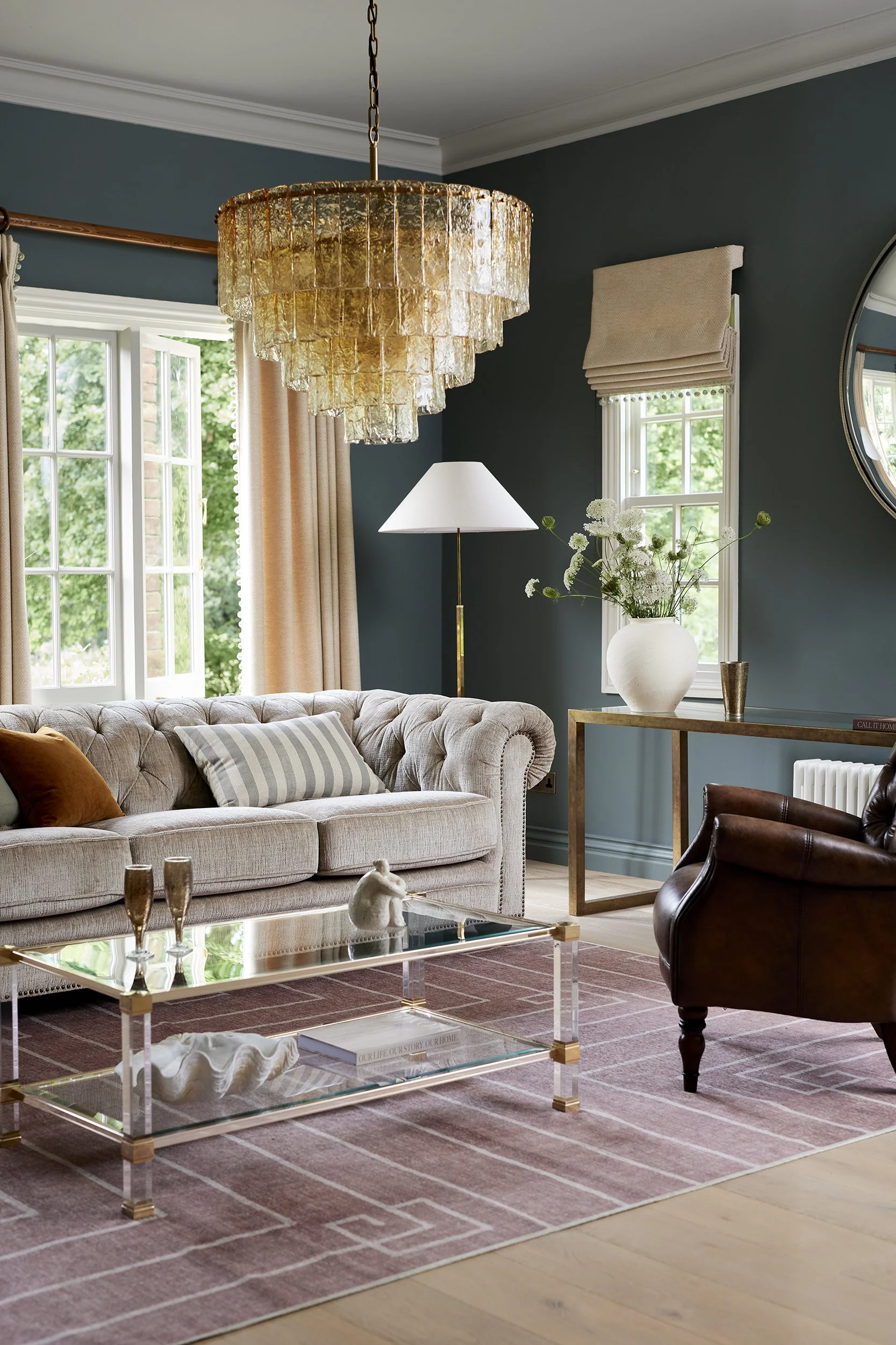 Living room with dark teal walls, beige tufted sofa with striped and orange pillows, glass coffee table with decor, leather armchair, gold side table with white vase and flowers, white floor lamp, large window with curtains, brass chandelier, mirror 