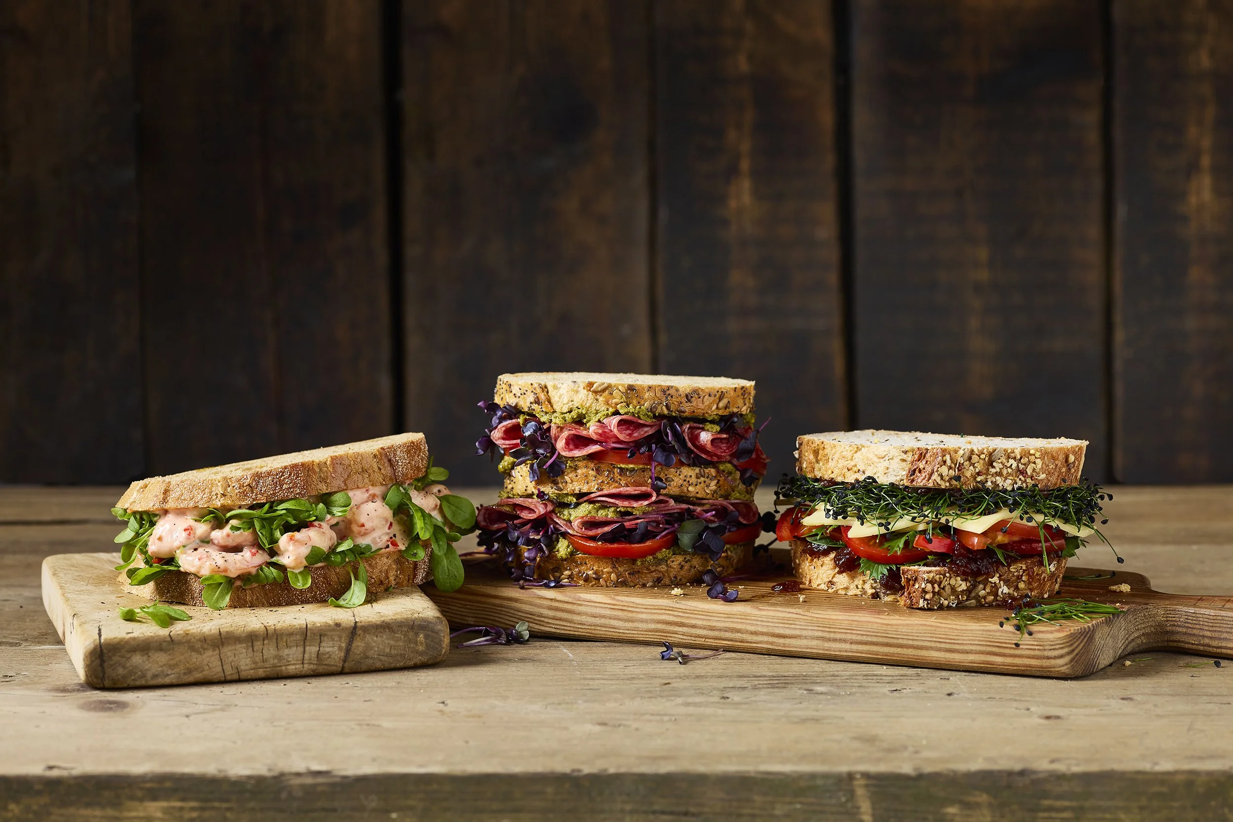 Three sandwiches with various ingredients on wooden boards against a dark wooden background.