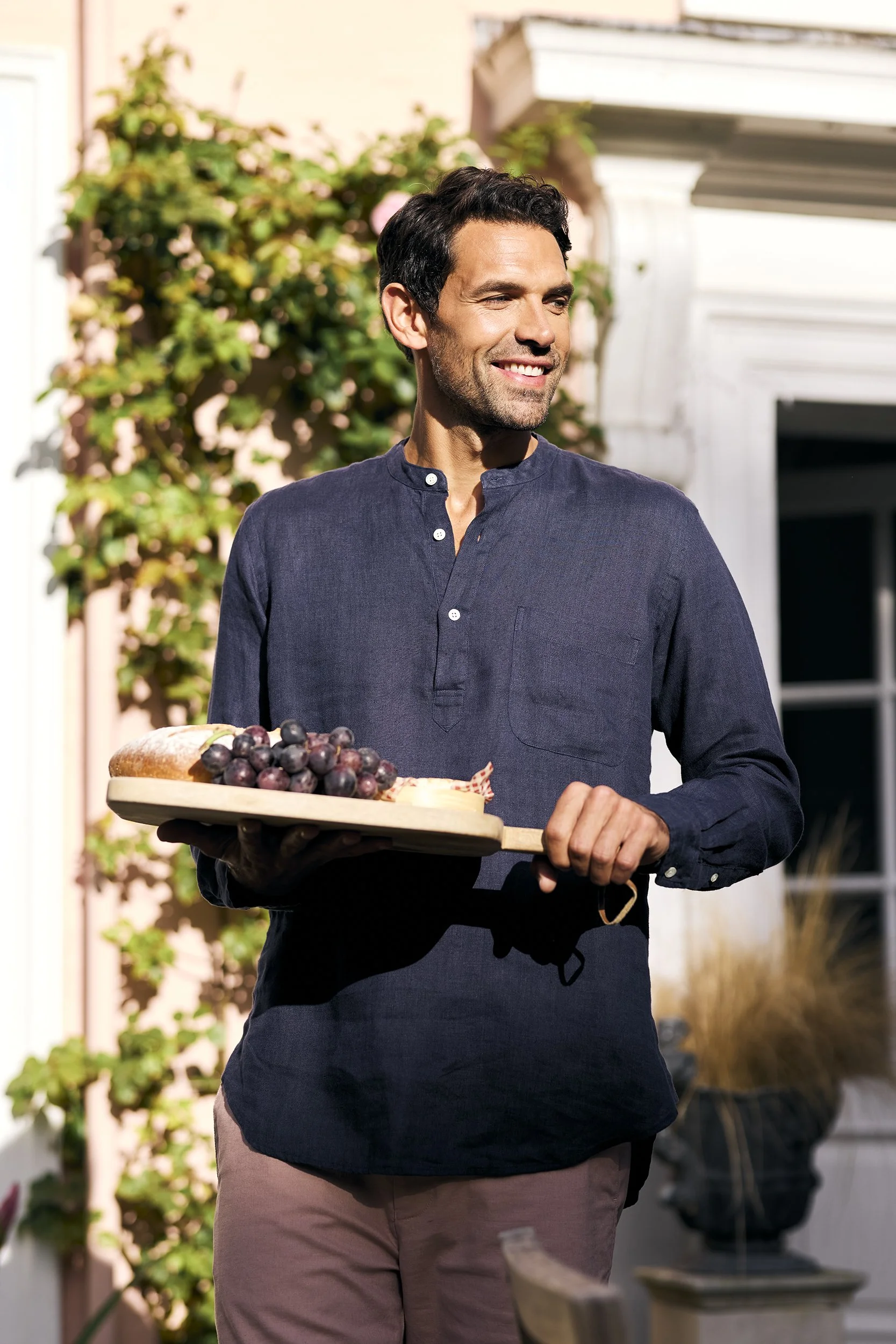 A man with dark hair and a beard smiling outdoors, holding a wooden tray with grapes and bread, in a backyard with a house and plants in the background.