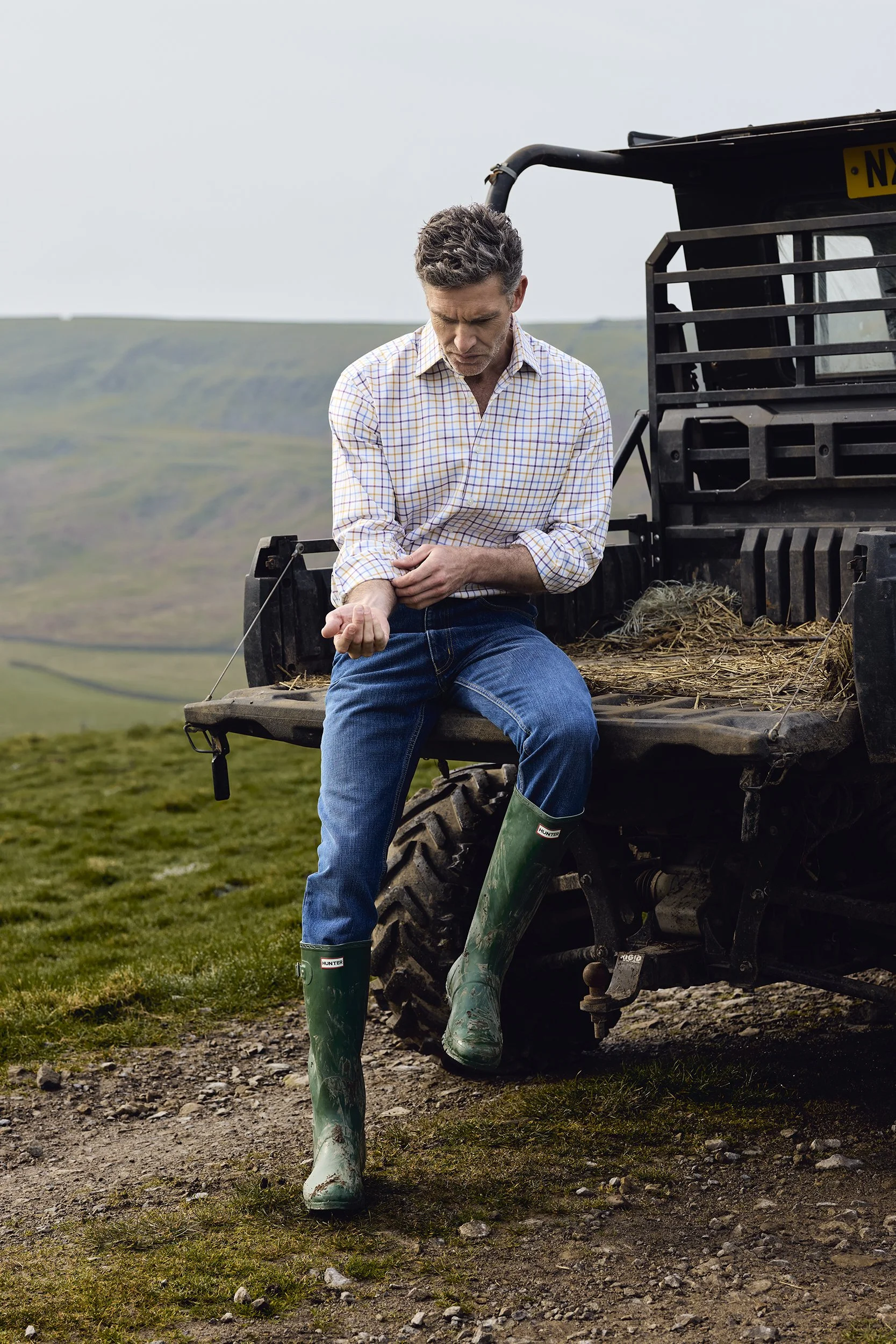A man sitting on the back of a utility vehicle, wearing green rubber boots and a plaid shirt, examining his arm in an outdoor rural landscape.
