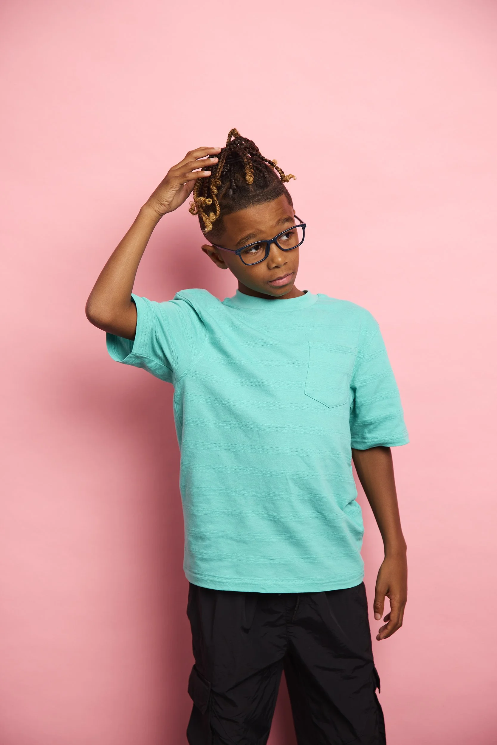 A young boy with glasses, wearing a turquoise t-shirt and black pants, standing against a pink background, scratching his head and looking to the side with a puzzled expression.