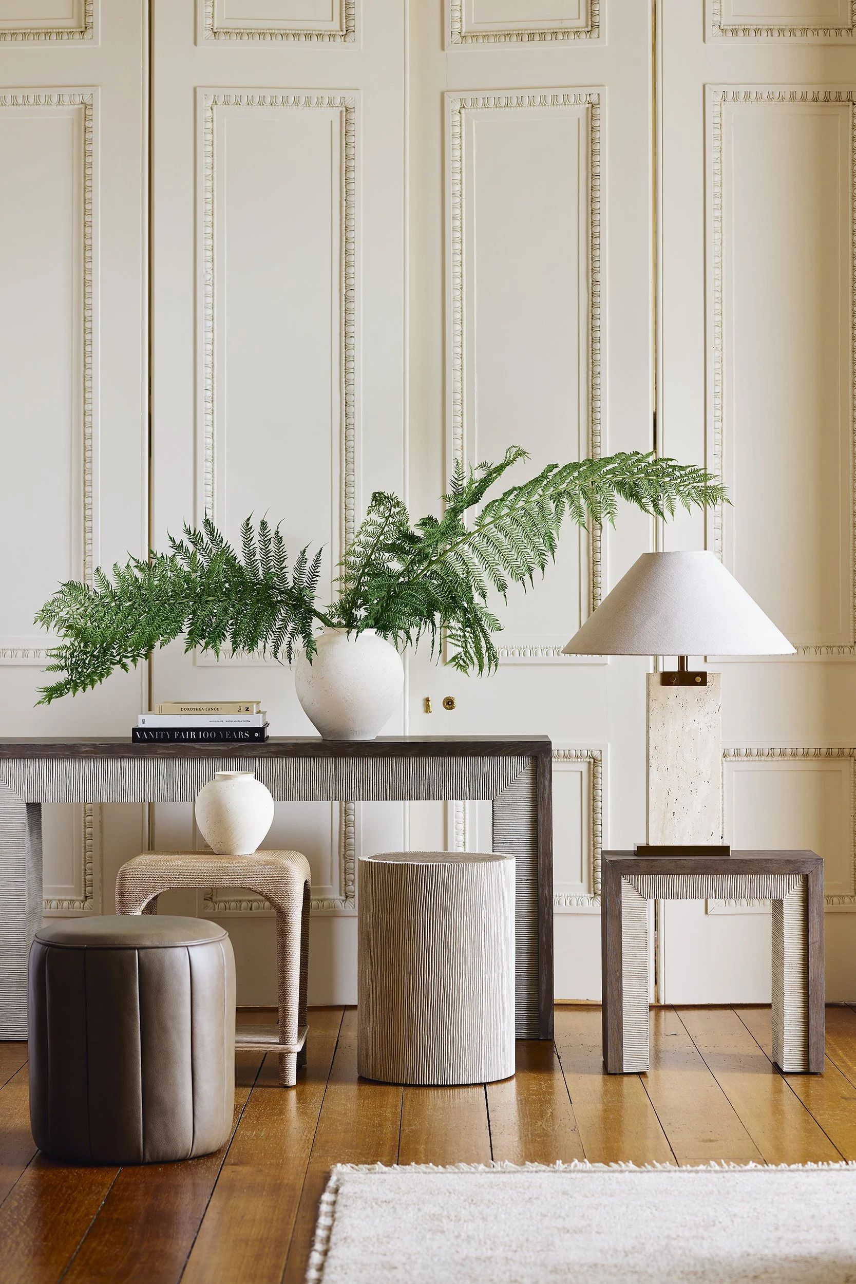 Interior scene with a wooden sideboard, green fern plant in a white vase, small decorative white vases, books, a table lamp, and a beige ottoman on a wooden floor with an area rug.