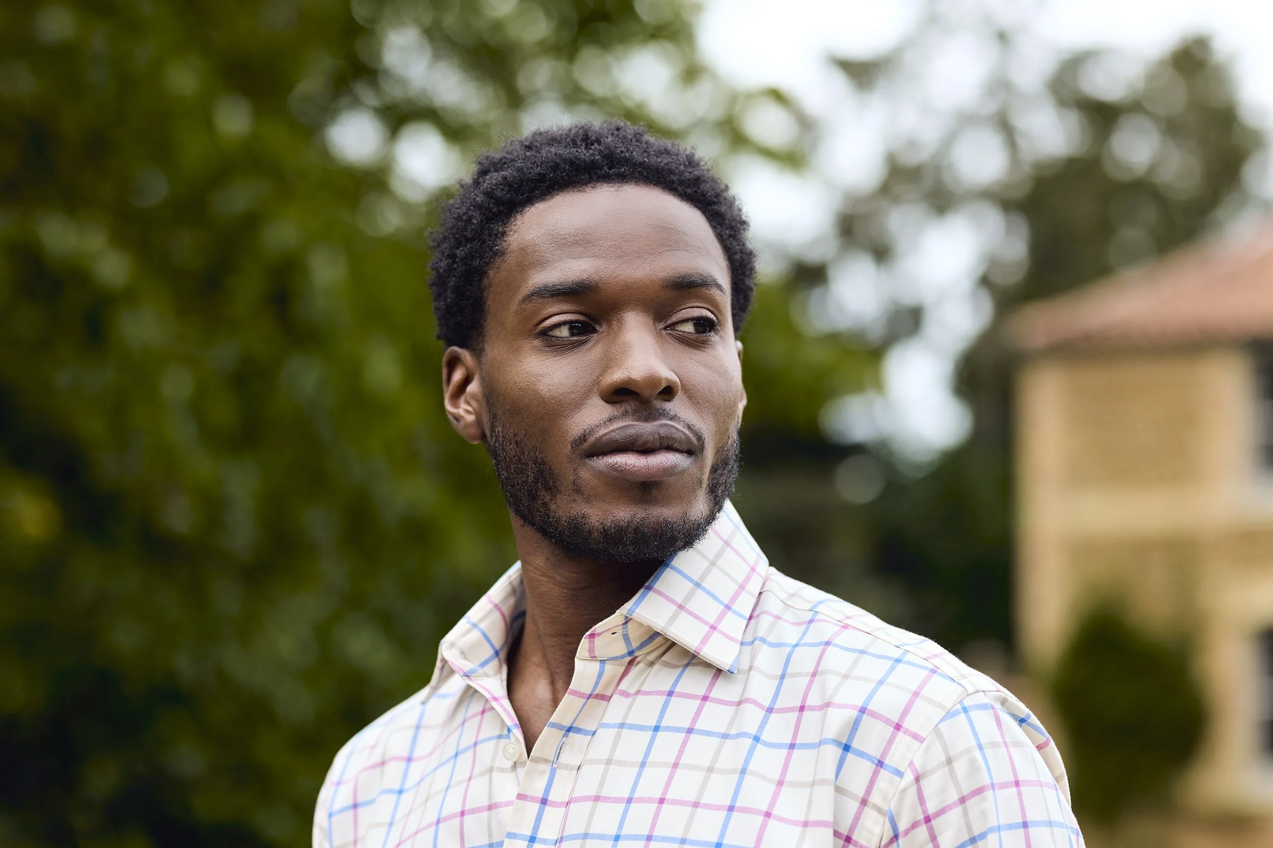 A young man with dark curly hair and a beard outdoors, looking to the side, wearing a checkered shirt.