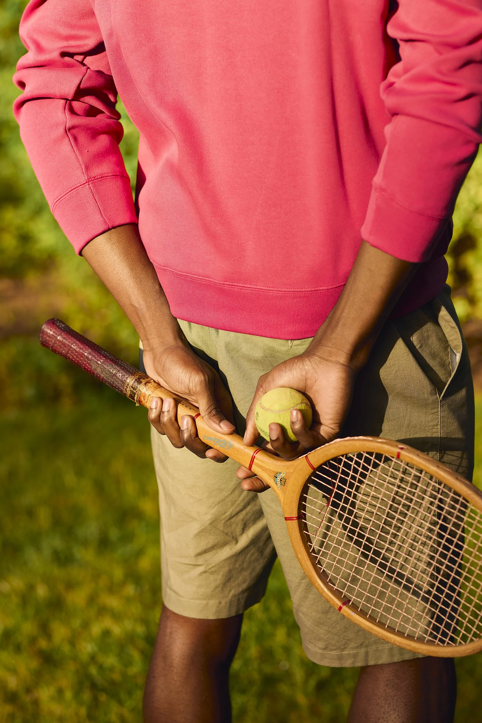 Person holding a tennis racket and tennis ball, wearing a pink sweatshirt and khaki shorts, outdoors on grass.
