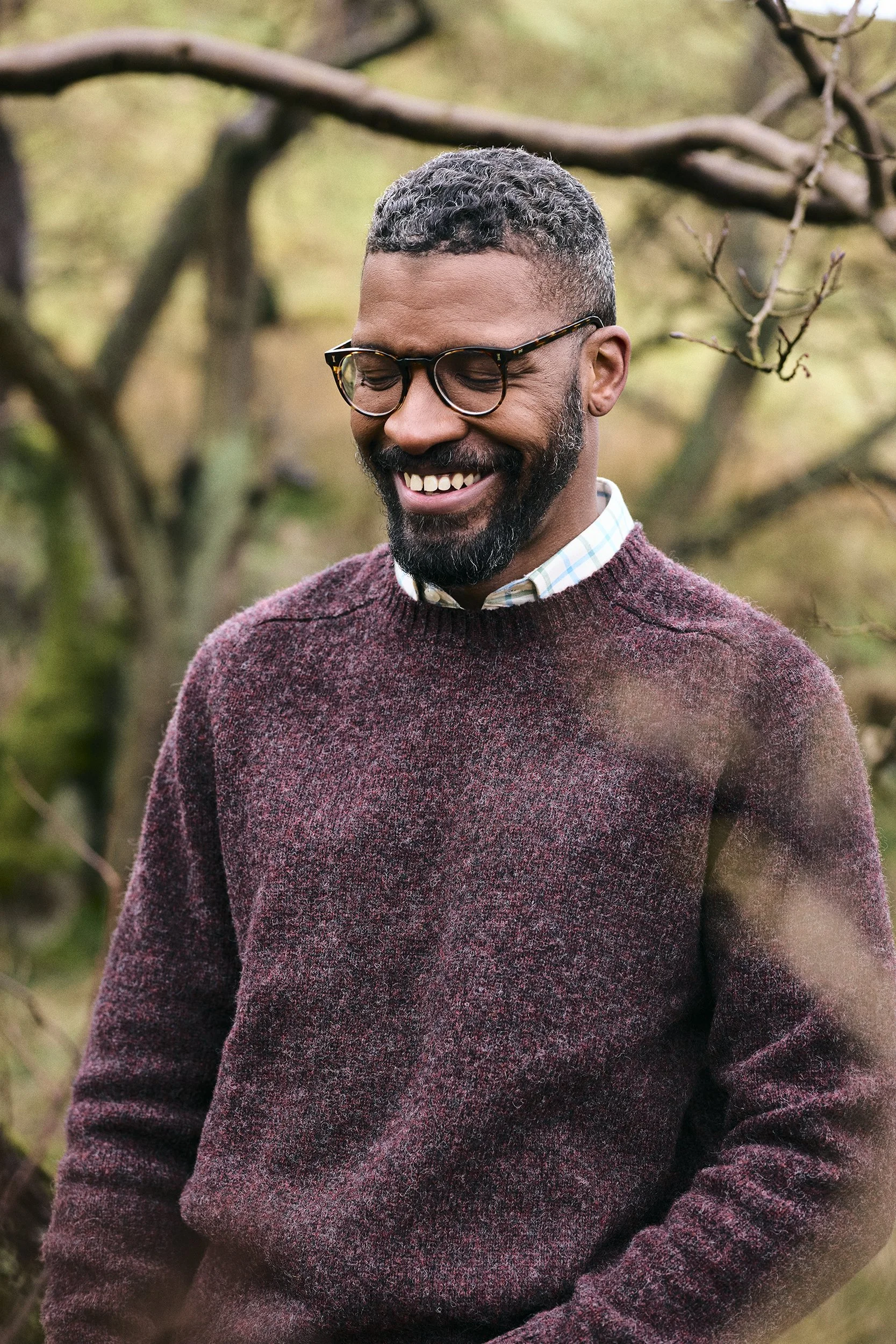 A smiling man with glasses and a beard, wearing a maroon sweater over a plaid shirt, standing outdoors with trees in the background.