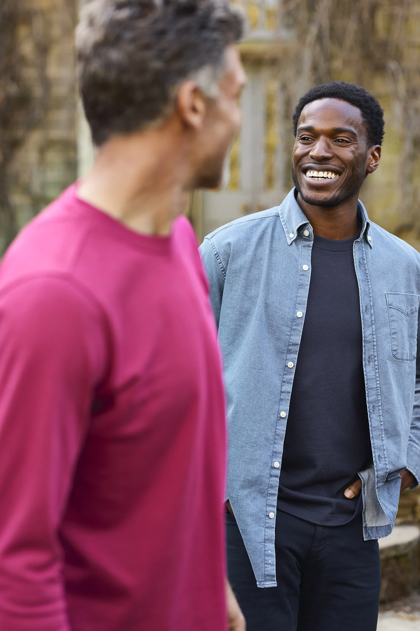 Two men are smiling and talking outdoors, with one wearing a pink shirt and the other in a blue denim shirt, in a yard with a wooden fence and autumn trees in the background.