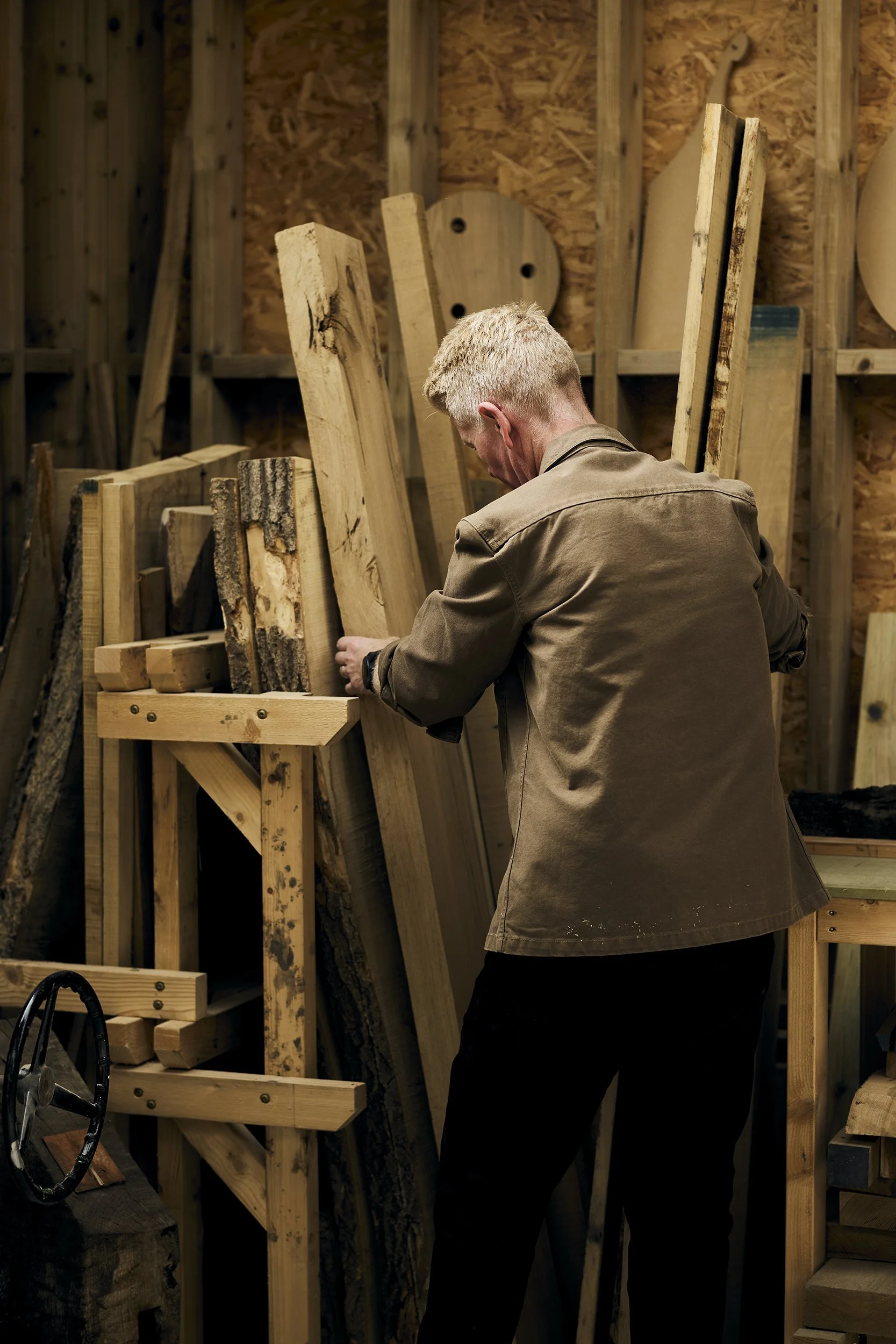 A man working in a woodworking shop, standing among wooden planks and pieces of lumber, examining a piece of wood.