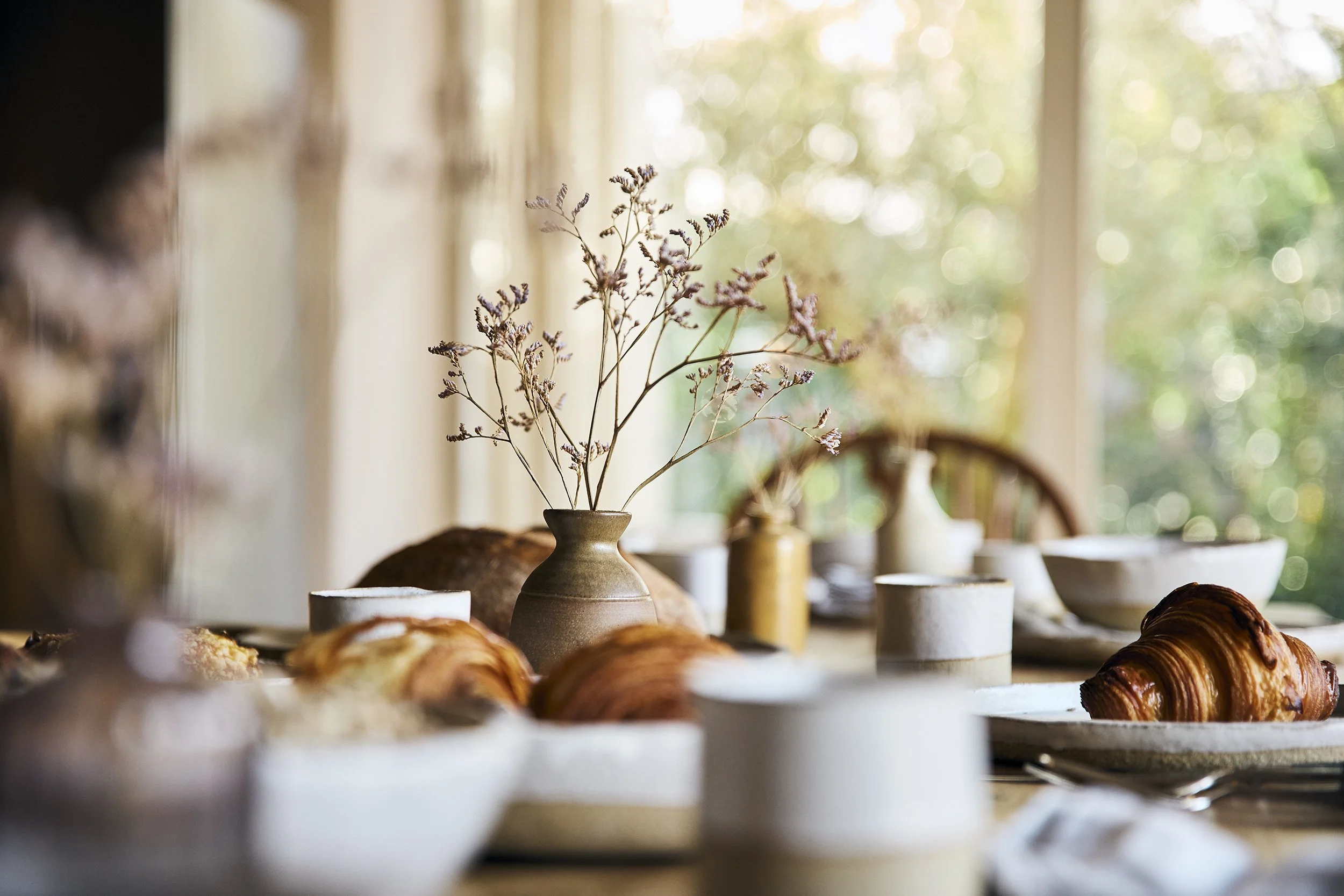 Breakfast table set with croissants, bread, and bowls, with a vase of dried flowers in the center, in a sunlit room.