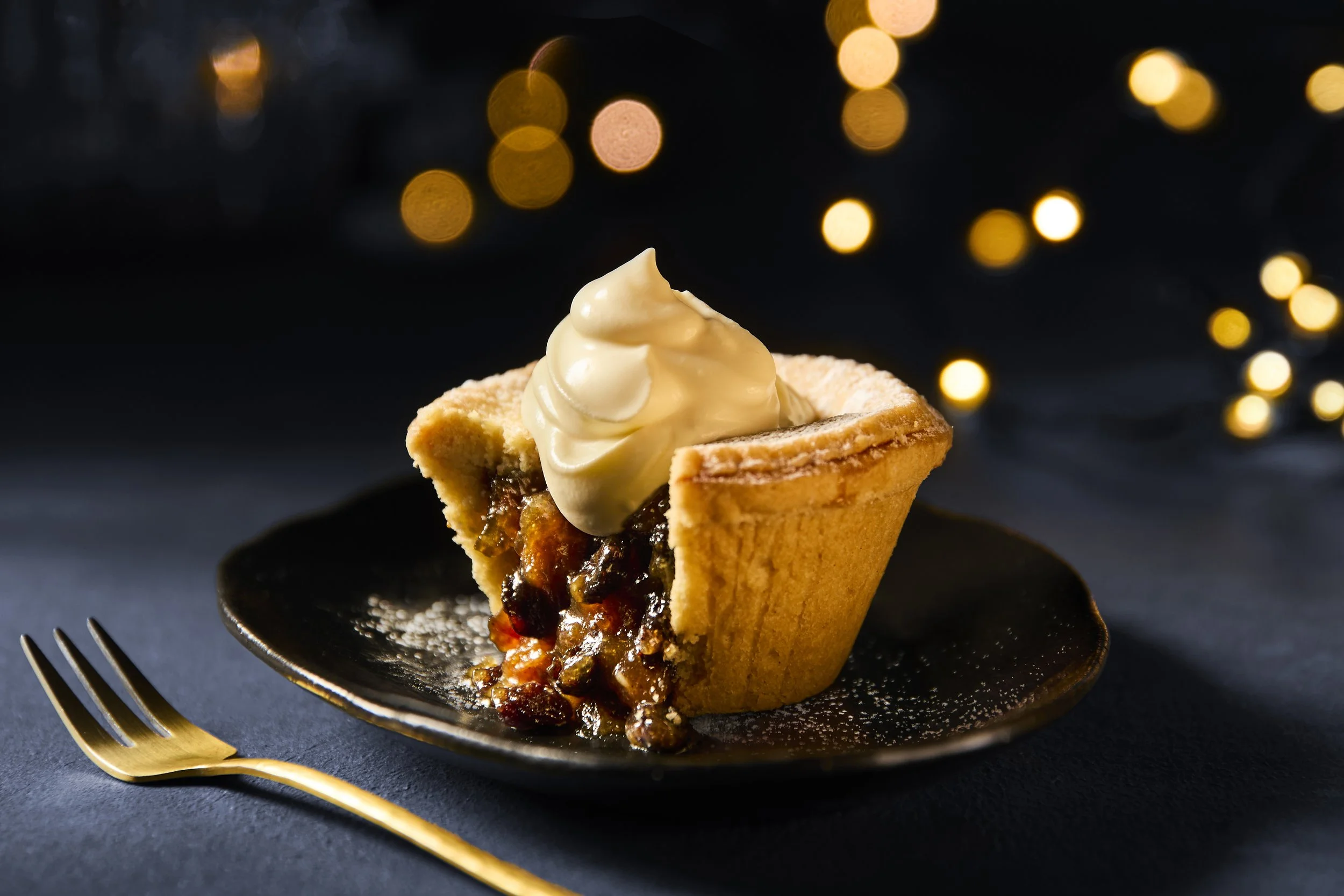 A slice of cherry pie with a dollop of whipped cream on top, served on a black plate with a gold fork, against a dark background with bokeh lights.