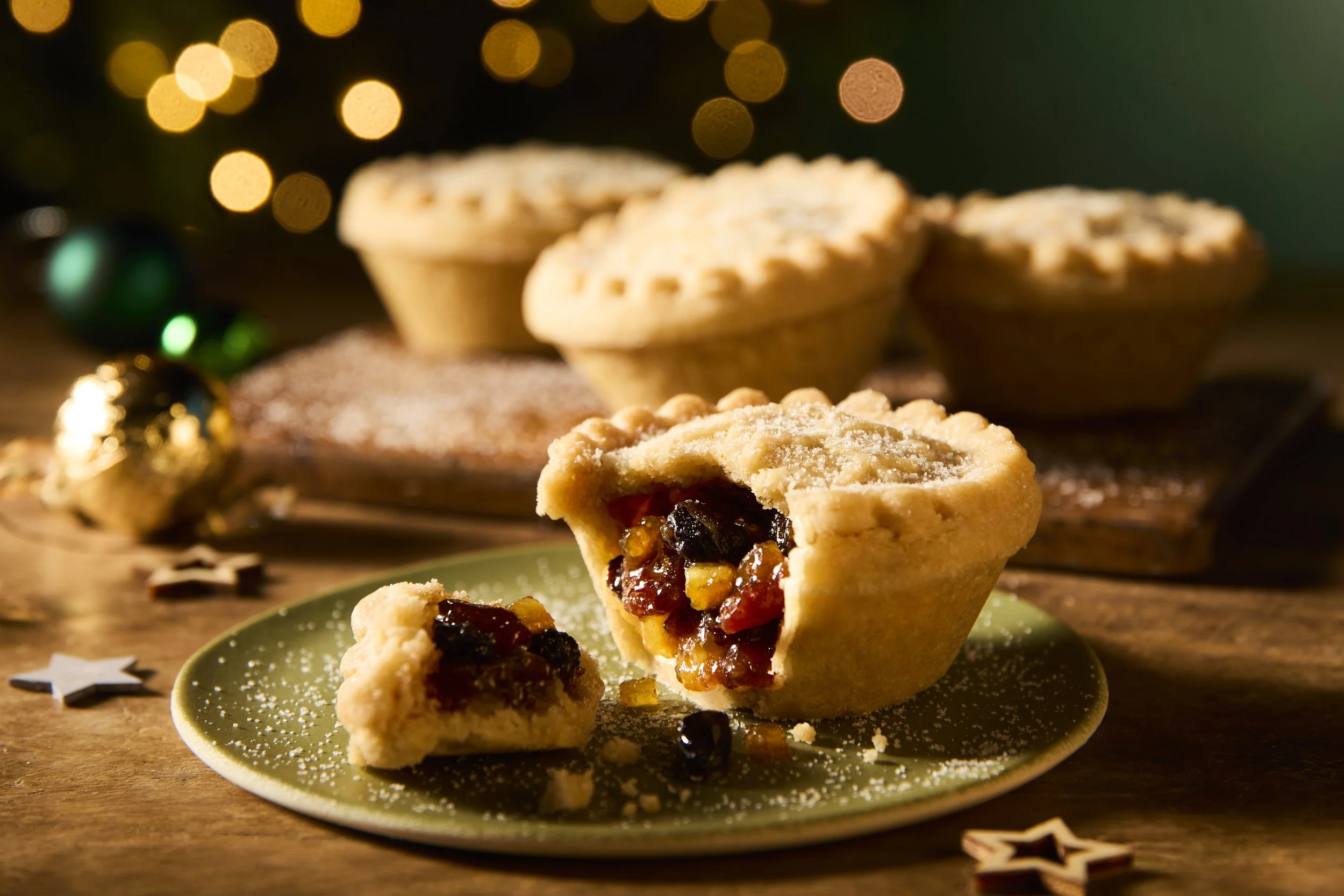 A partially eaten homemade fruit pie on a green plate with a flaky crust and a dark fruit filling, set on a wooden surface with holiday decorations and additional pies in the background.