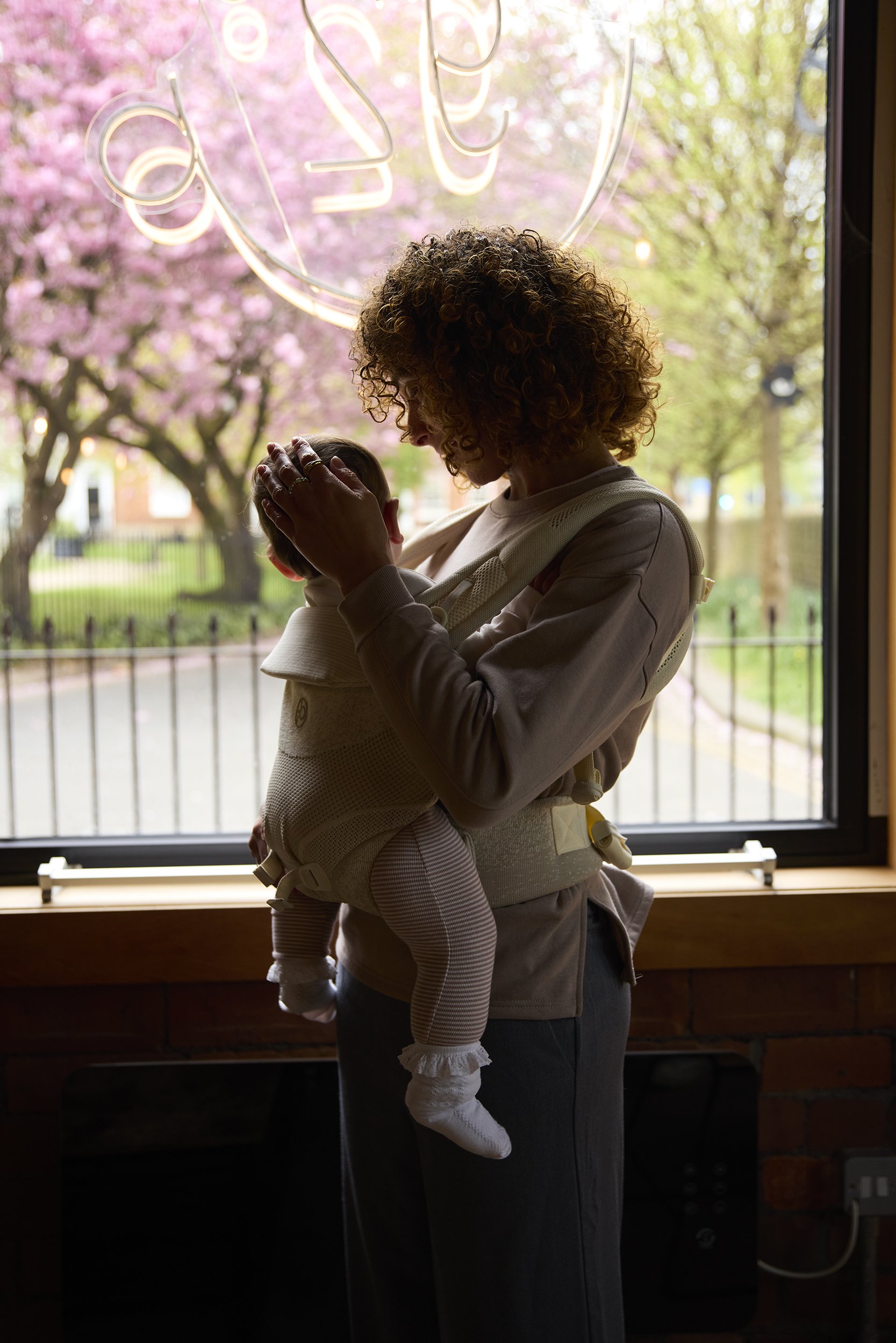 A woman with curly hair holding a baby near a window with pink blossoms outside.