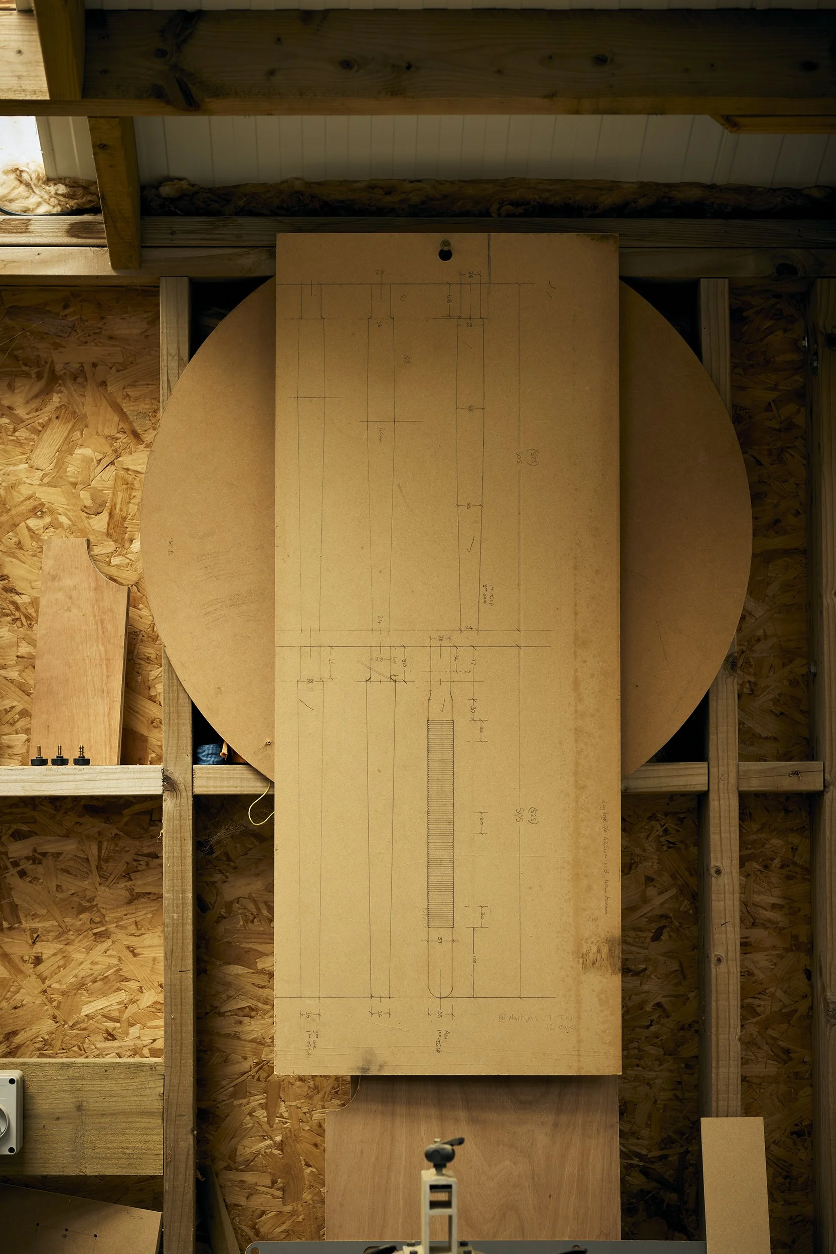 Hand-drawn technical blueprint on a large piece of wood, mounted on a workshop wall with wooden framing and insulation in the background.