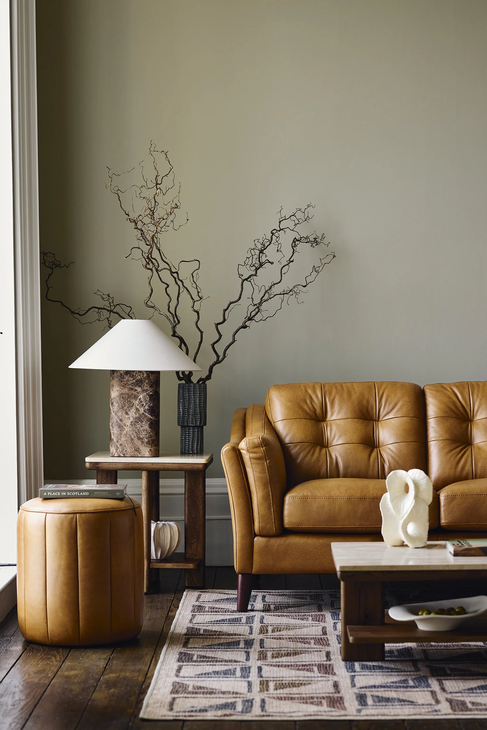Living room with brown leather sofa, wooden coffee table with decorative sculpture, patterned rug, side table with lamp, and dried branches in a vase against a green wall.