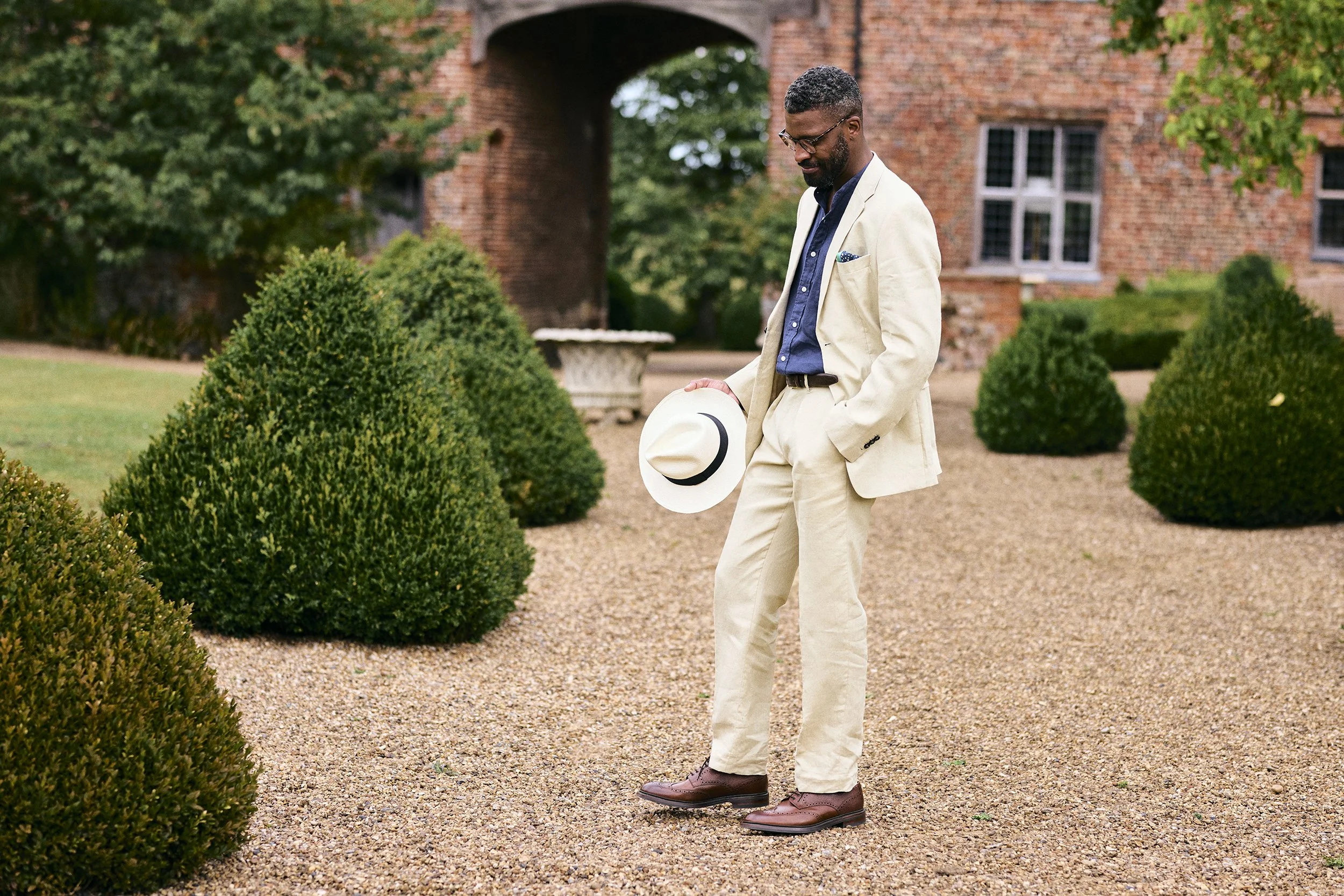 Man dressed in beige suit holding a white hat, standing on a gravel path surrounded by neatly trimmed bushes and a brick building in the background.