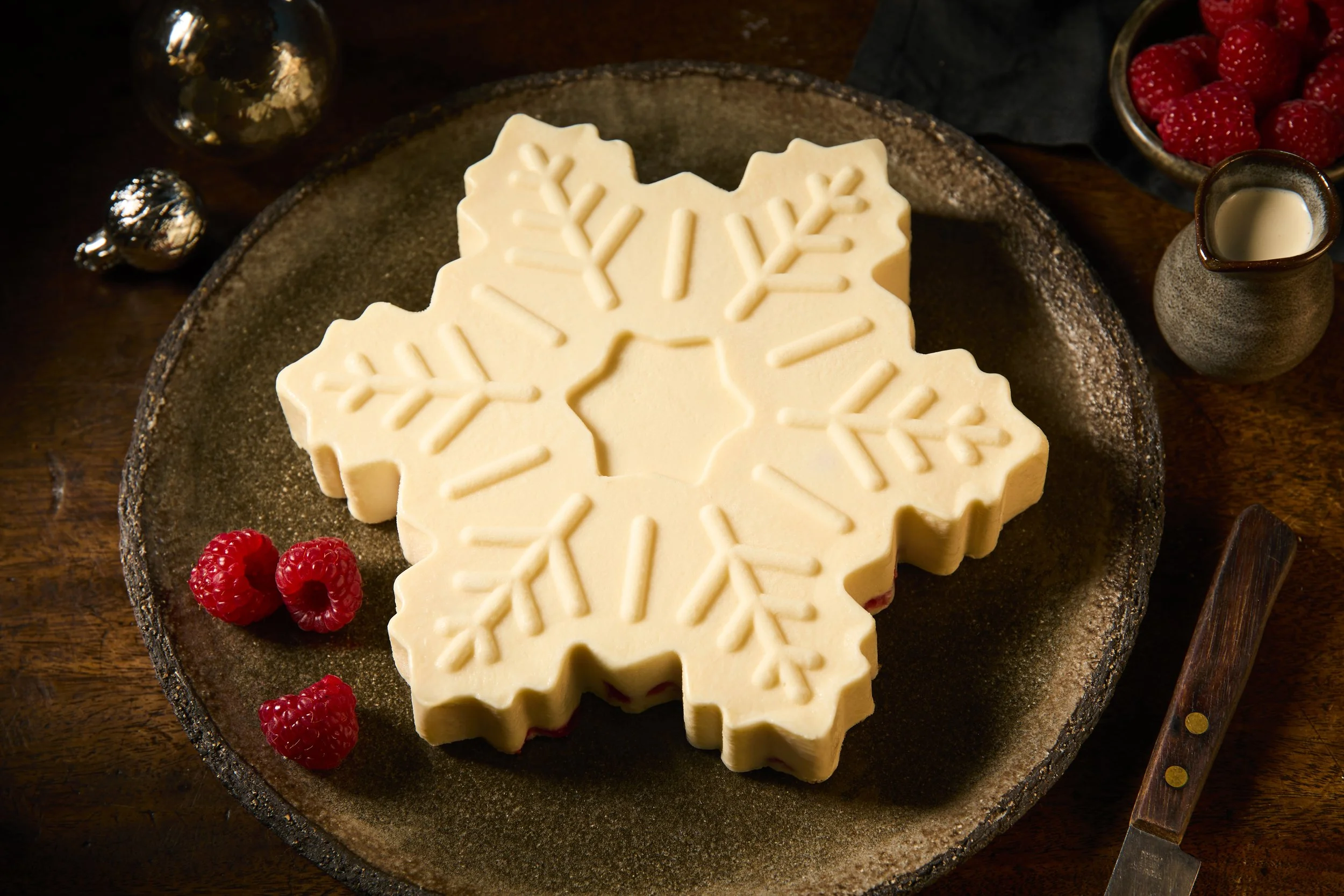 A snowflake-shaped white chocolate bark on a dark round plate, garnished with fresh raspberries, surrounded by small Christmas ornaments, a small jug of white chocolate, and a wooden knife on a wooden table.