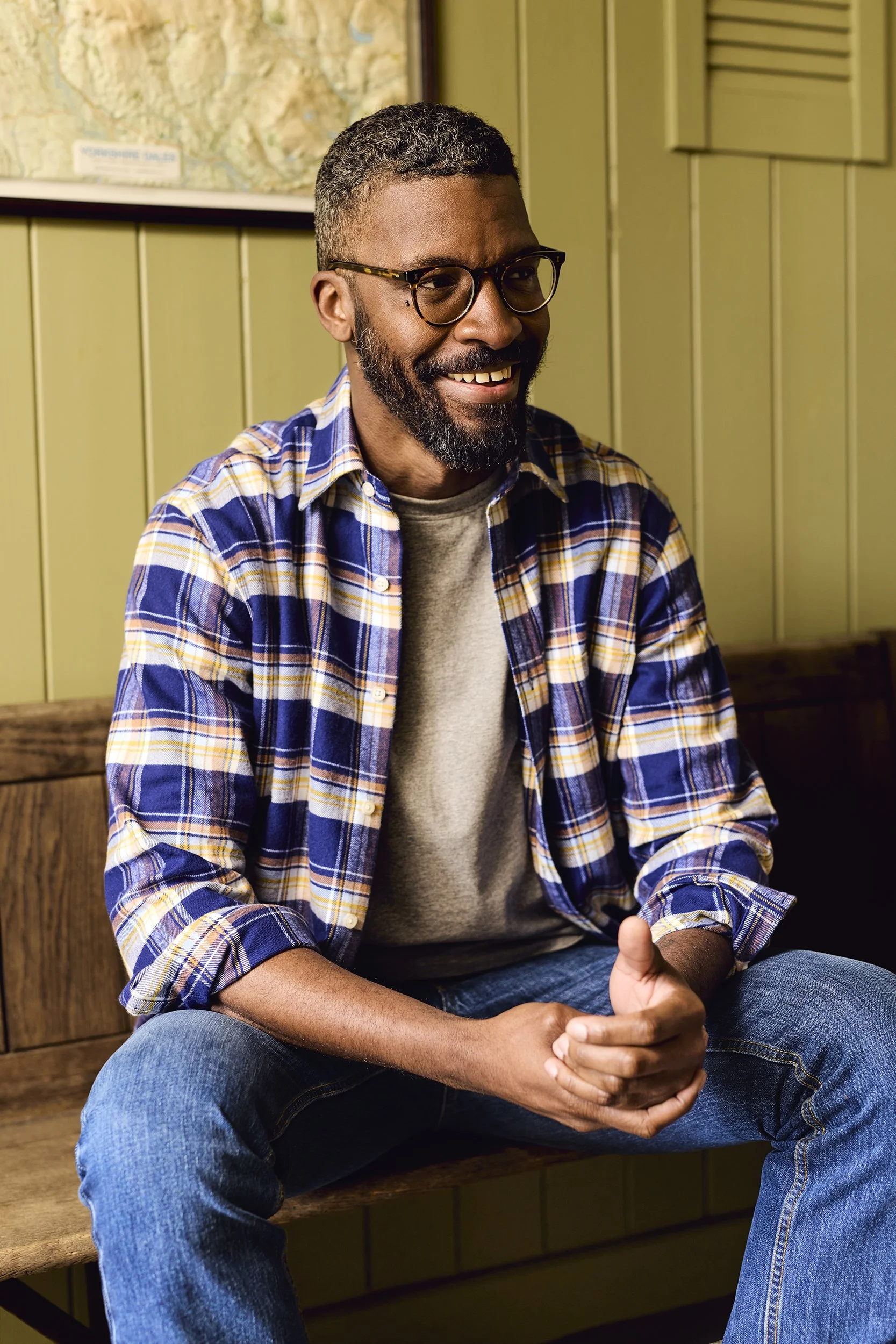 A man with glasses, a beard, and curly hair, smiling and sitting on a wooden bench, wearing a plaid shirt over a gray t-shirt, in a room with green wooden panel walls and a map on the wall.