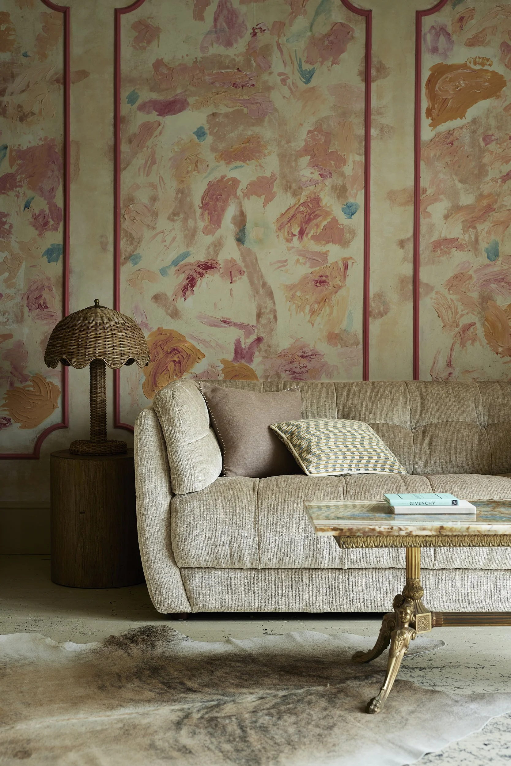 A beige sofa with pillows, a wooden side table with a wicker lamp, a patterned rug, and an ornate side table in a living room with a colorful abstract floral wall mural.