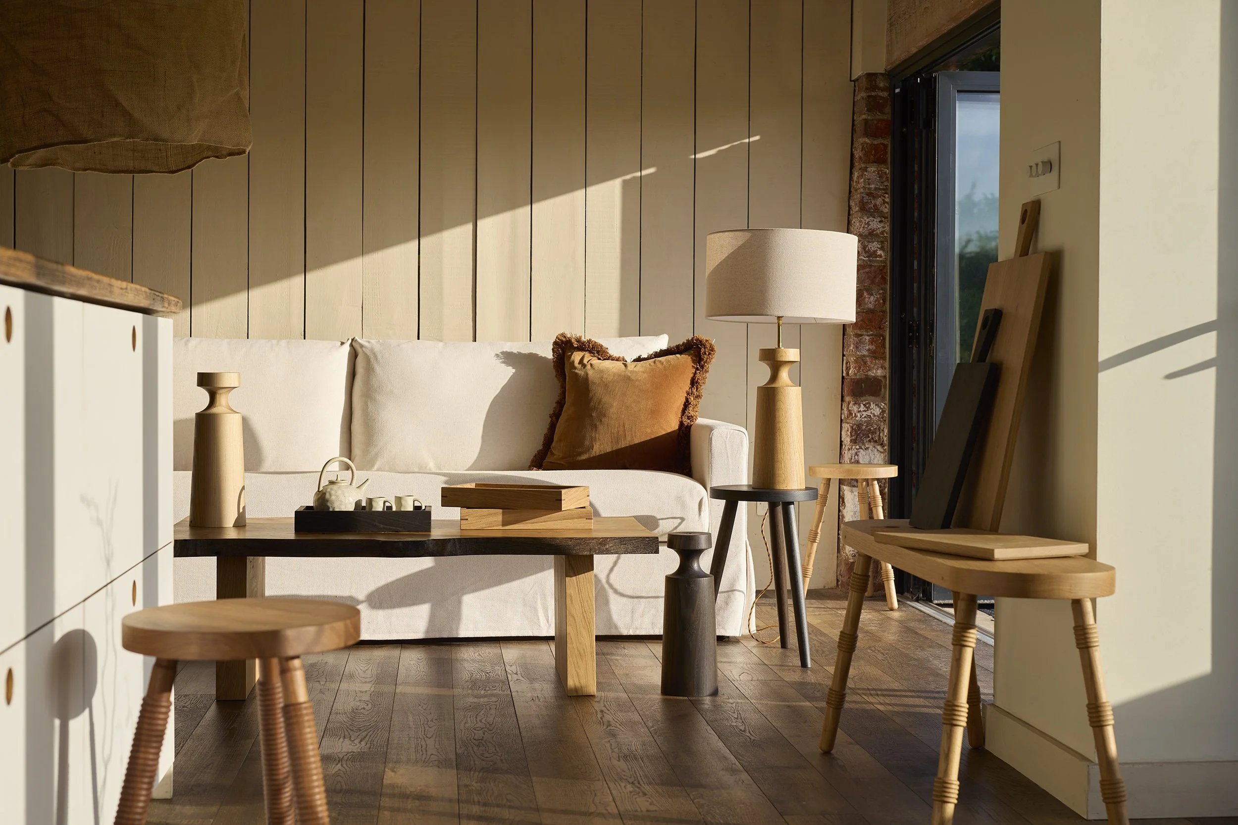 Cozy living room with a white sofa, wood panel wall, and natural light streaming through a door with black trim. Coffee table and stools made of wood, decorative pillows, and lamps add to warm, earthy decor.