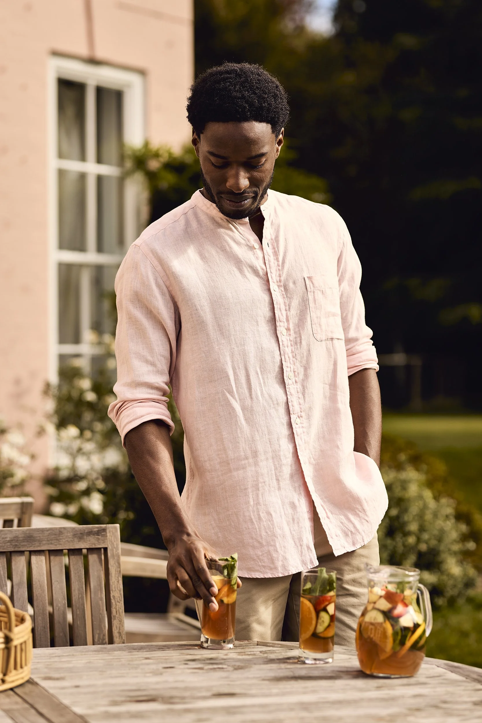 A man with dark skin, black curly hair, wearing a light pink shirt, standing outdoors at a patio table with a pitcher and two glasses of fruit-infused iced tea.