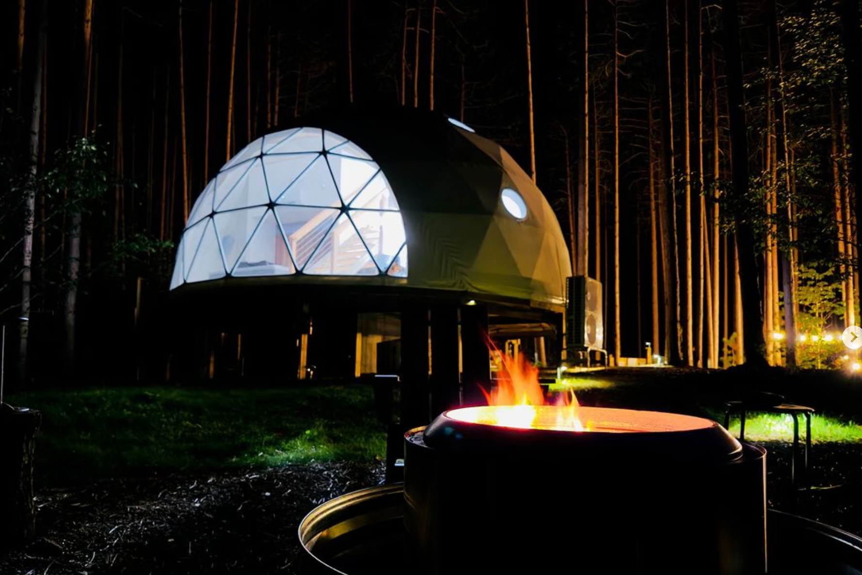 A geodesic dome-shaped cabin illuminated at night in a forest setting with tall trees, a campfire in the foreground, and warm outdoor lighting.