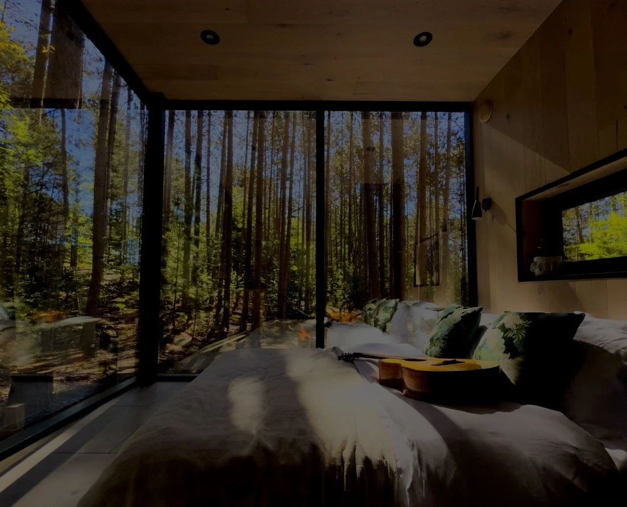 Interior of a modern bedroom with large glass windows showing a forest outside. The bed has white linens, several pillows with green floral patterns, a wooden guitar, and a small black object on it. The room features wooden walls and ceiling, with some black lighting fixtures and a small window with a decorative plant inside.