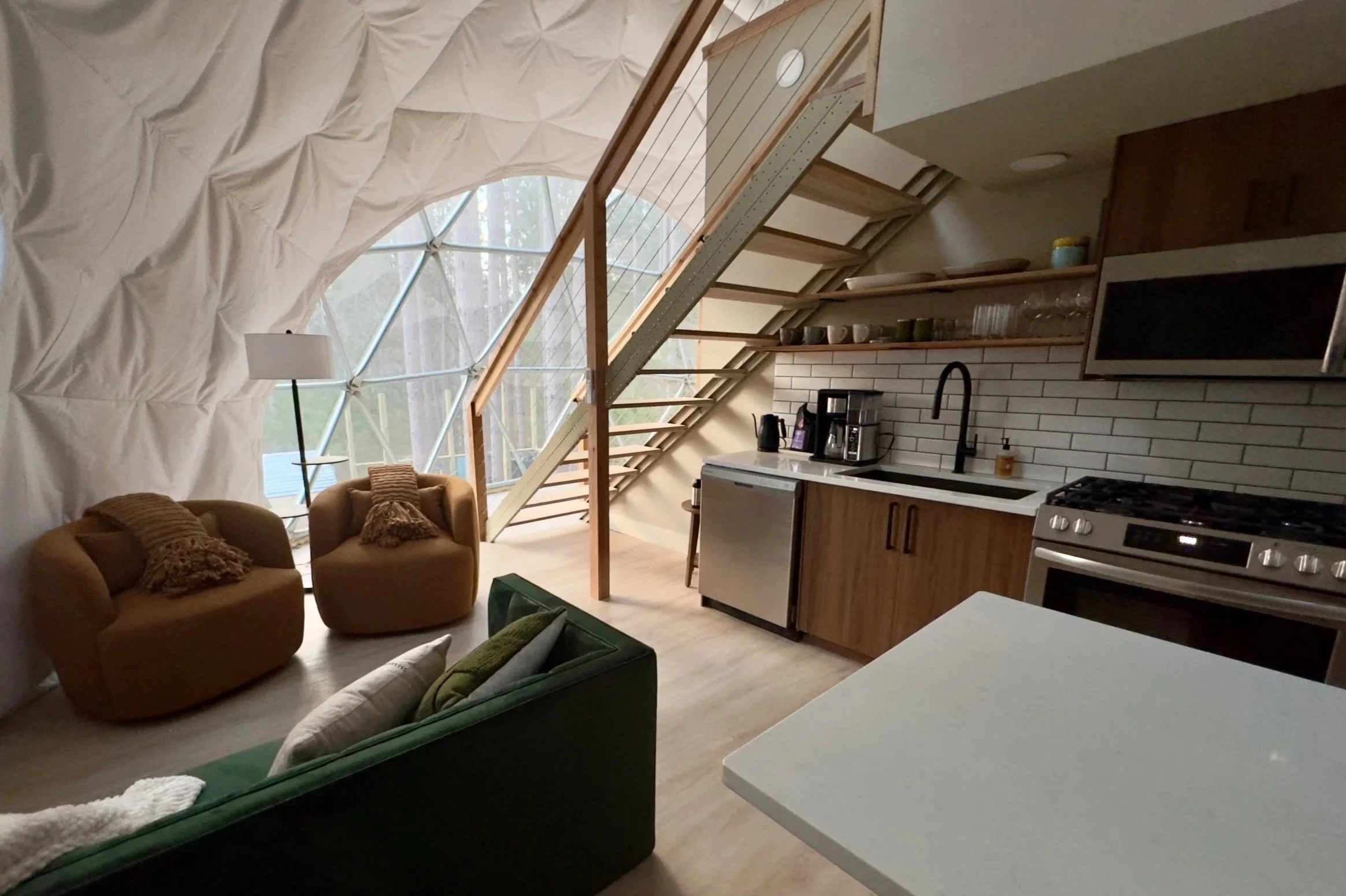 Interior of a modern home with a high ceiling and large windows, including a living area with two armchairs, a sofa, and a kitchen with open shelving, a stove, and a countertop.