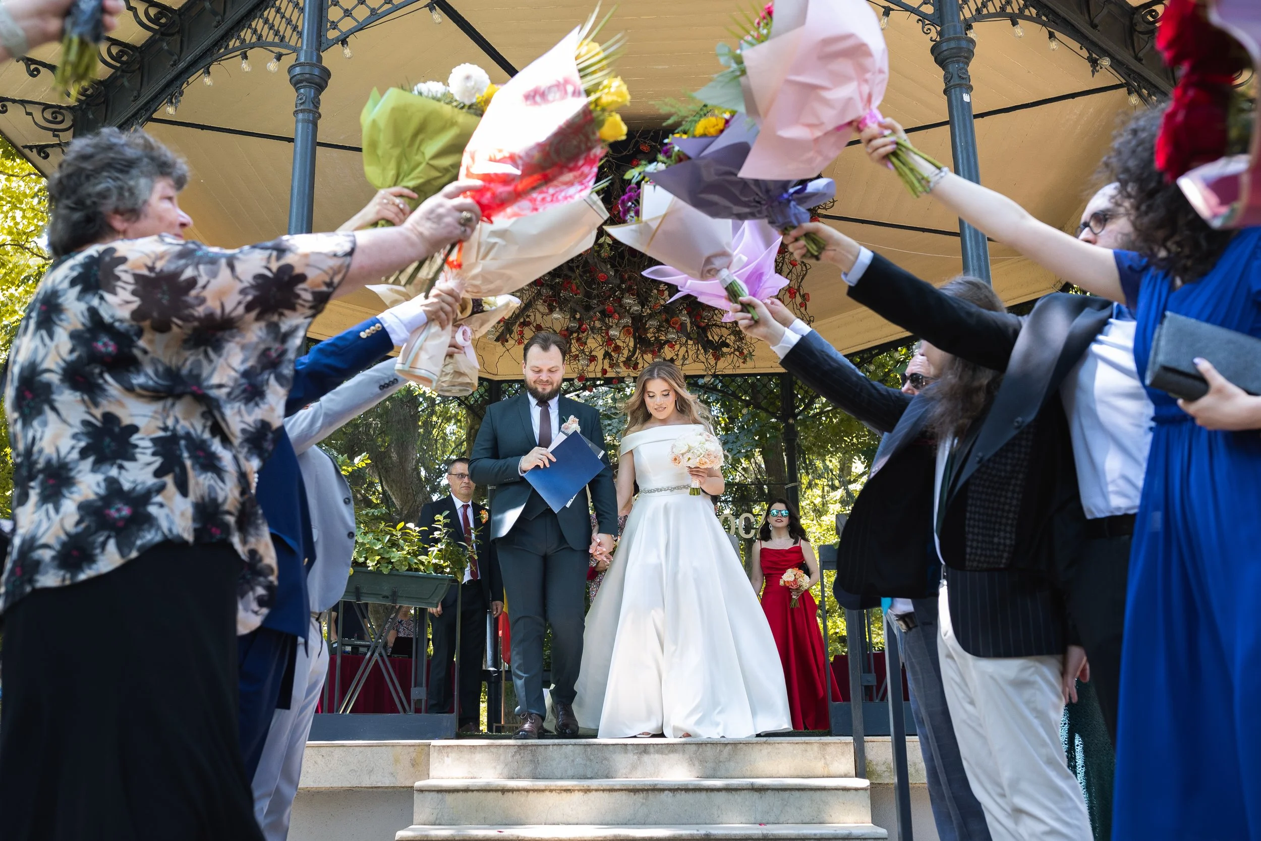 A bride and groom walking down the steps of a gazebo, holding hands, as their friends and family surrounding them celebrate and shower them with bouquets of flowers during an outdoor wedding ceremony.