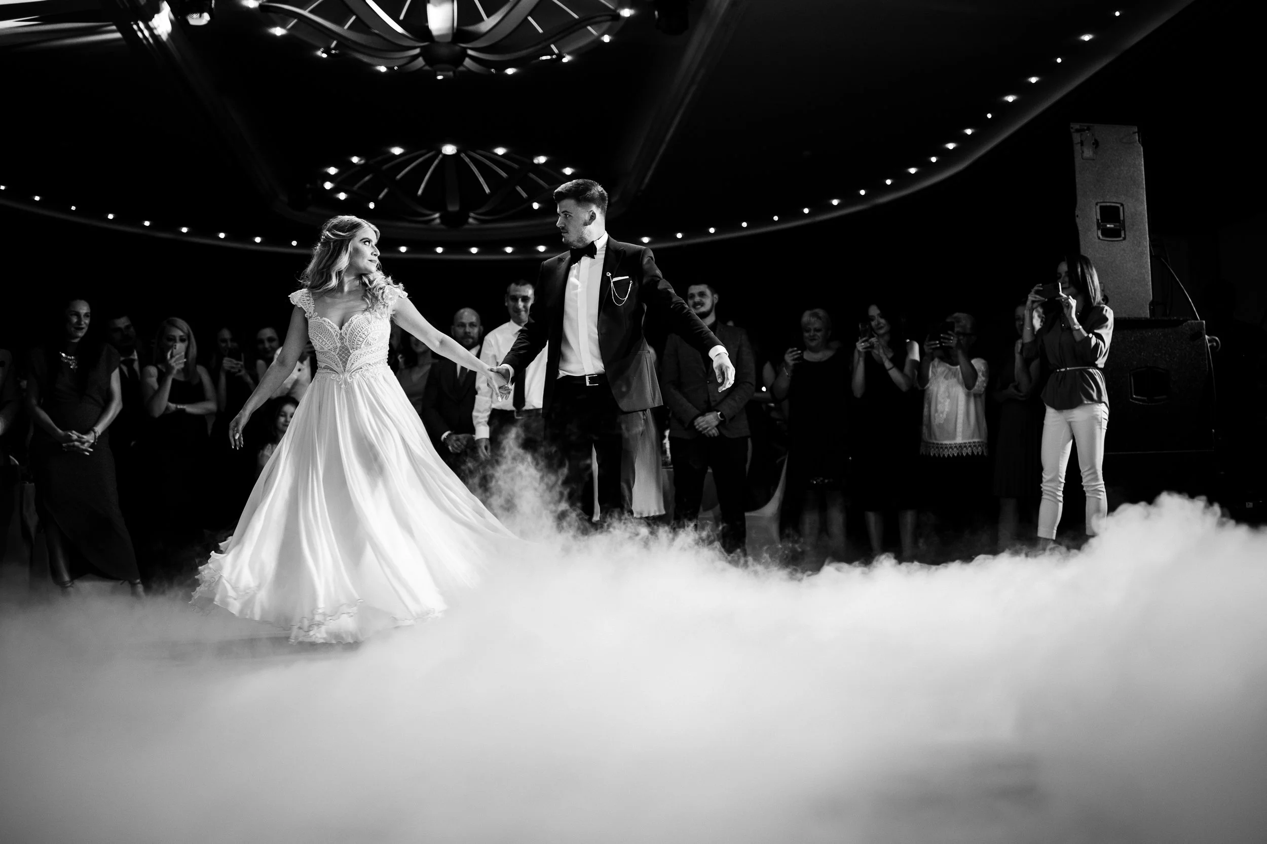 A bride and groom dancing at their wedding reception, surrounded by guests, with a smoky dance floor and overhead lighting.