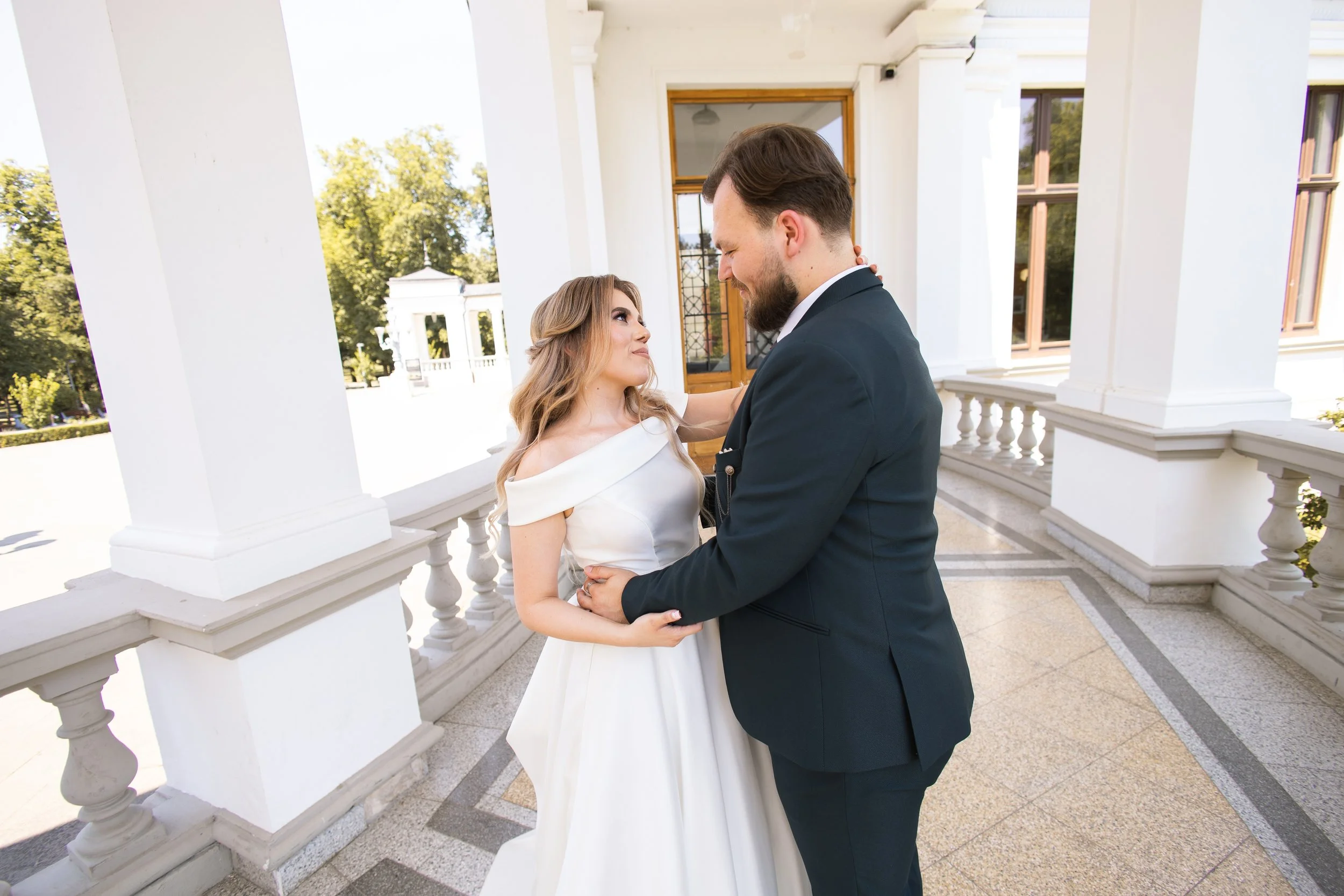 A bride and groom standing close together on a white porch, looking into each other's eyes, with a garden and trees in the background.