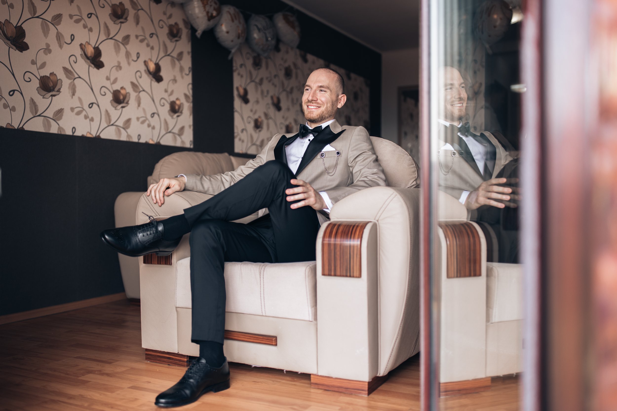 A young man in a tuxedo with a beige blazer and black bow tie is sitting on a cream-colored sofa, smiling and relaxed, in a room with floral wallpaper.