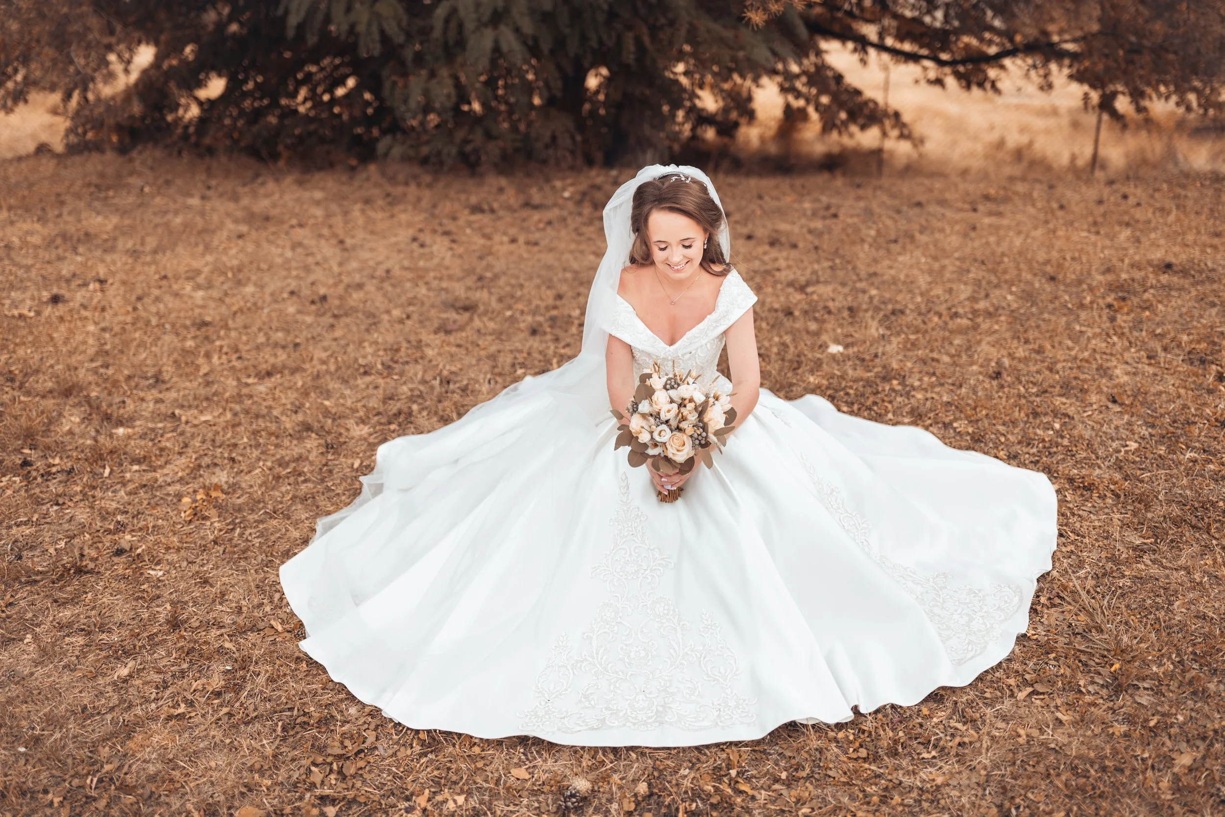 A bride in a white wedding dress sitting on the ground outdoors, holding a bouquet of flowers, with trees and dry grass in the background.